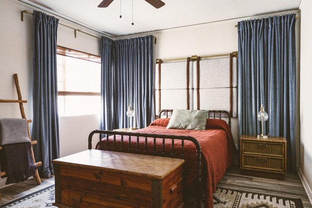Bedroom with a metal bed frame, red bedding, green pillows, wooden nightstand with lamps, blue curtains, a window, ceiling fan, and a wooden storage chest at the foot of the bed.
