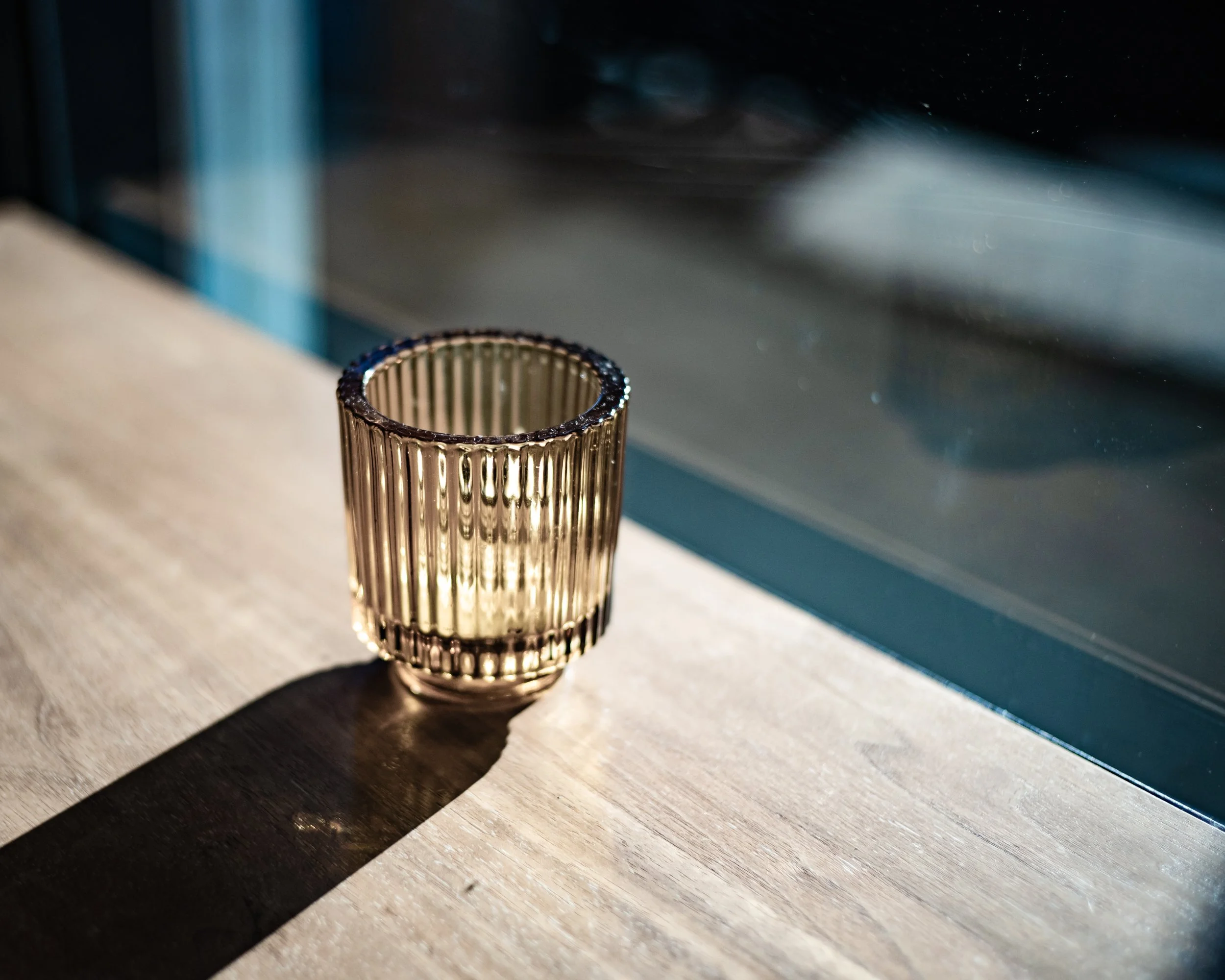 A gold-colored glass candle holder with a ribbed design, casting a shadow on a wooden surface near a window.