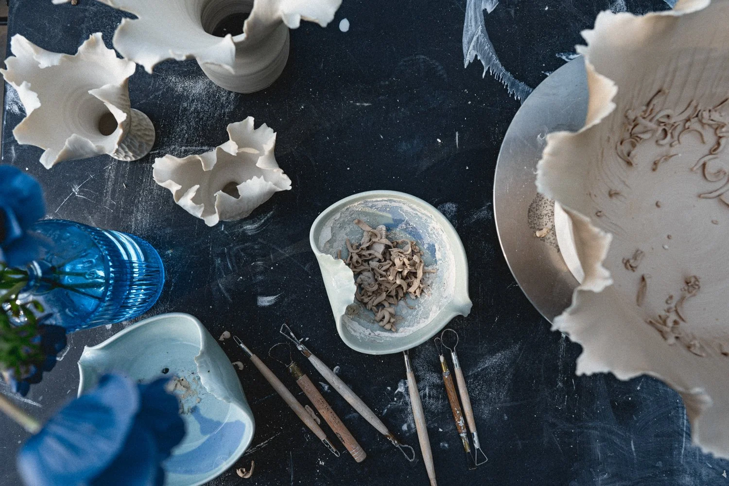 Workstation with sculpting tools, broken ceramic pieces, a vase, and a partly-finished ceramic sculpture on a dark table.
