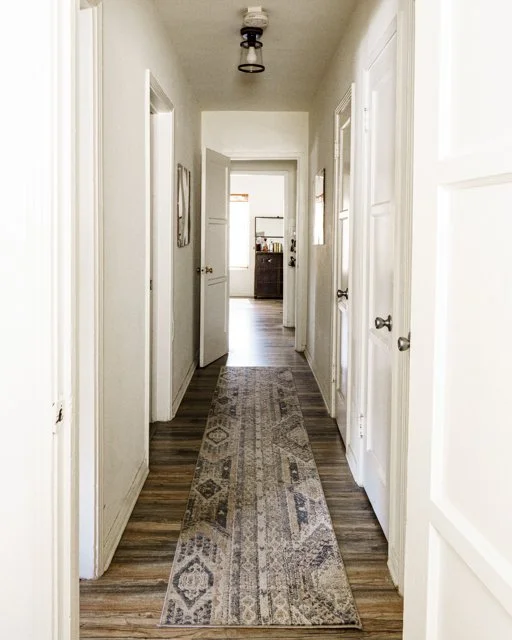 A hallway with white walls and six doors, three on each side. A patterned runner rug runs down the center of wood floor, leading to a room at the end with a kitchen visible in the background.
