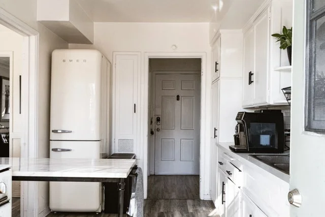 Kitchen with white cabinets, a vintage cream-colored refrigerator, and a black coffee maker on the counter. The front door is visible in the background.