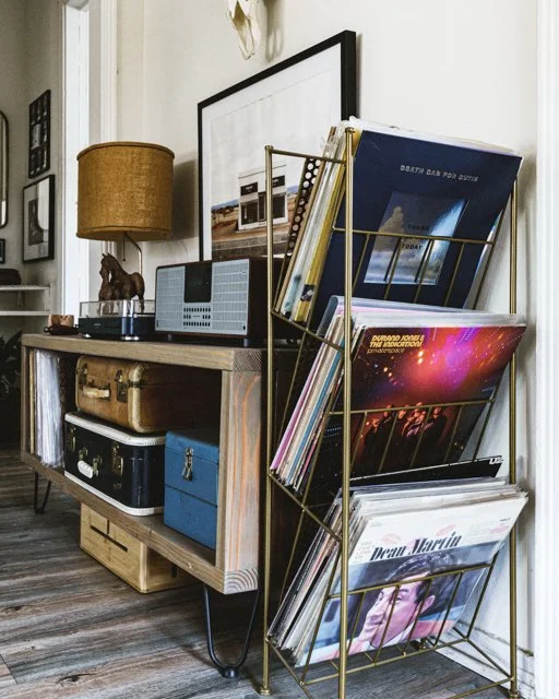 Side view of a living room corner with a wooden console table holding a lamp, a radio, and decorative items, next to a gold magazine rack filled with vinyl records and magazines.