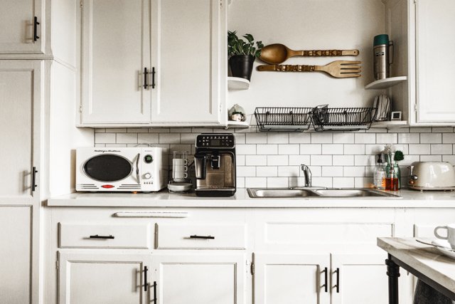 Kitchen counter with a microwave, coffee maker, dish rack, toaster, and utensils. White cabinets and subway tile backsplash.