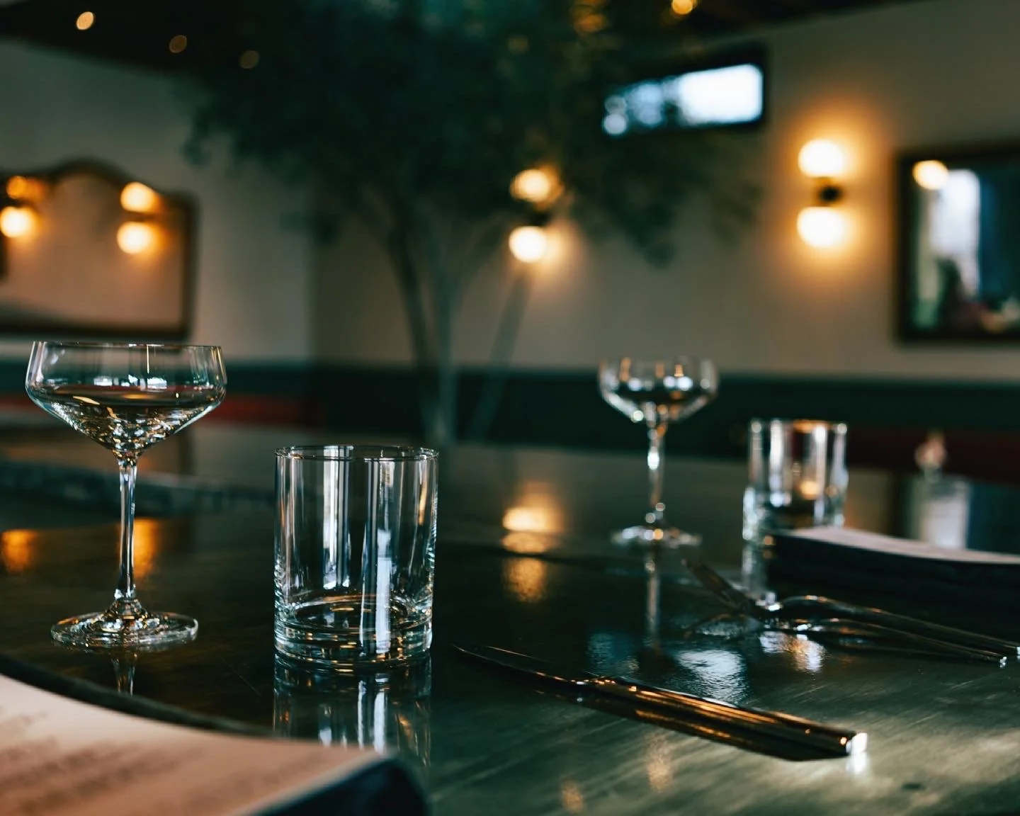 Empty restaurant table with wine glasses, water glasses, silverware, and napkins in a dimly lit setting.