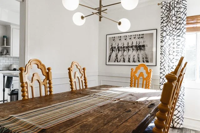 A dining room with a rustic wooden table and six wooden chairs with carved backs. A modern chandelier with multiple white globes hangs above the table. A black and white art print of a parade or marching band decorates the white wall. Natural light c