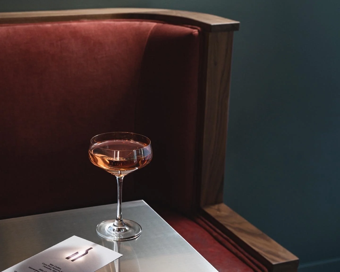 A glass of rosé wine on a table next to a white sheet of paper with a black border, in front of a red upholstered headboard and a dark colored wall.