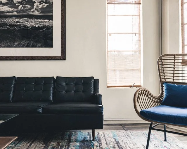 Living room with black leather sofa, wicker armchair with blue cushion, artwork on the wall, and a window with blinds.