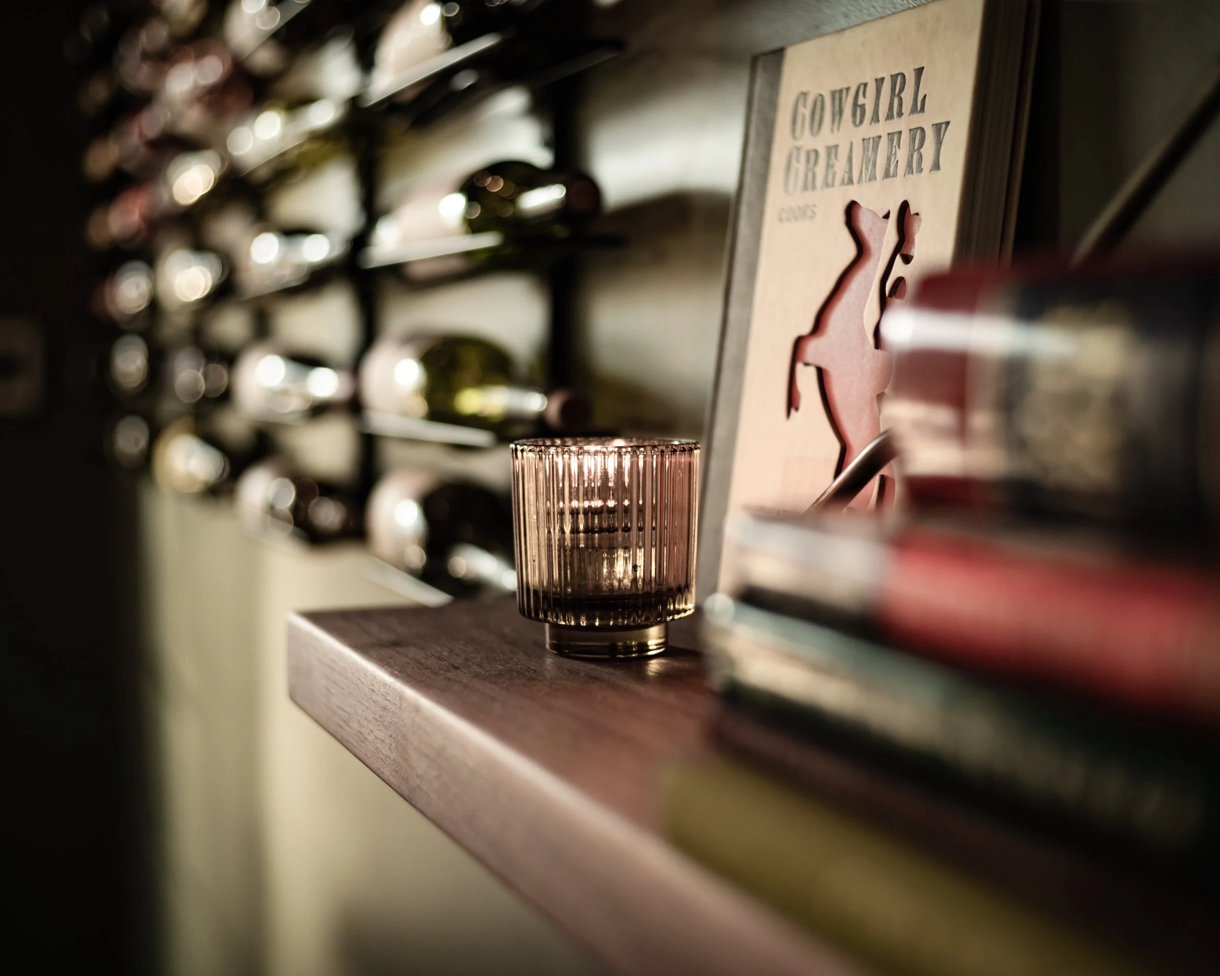 A glass with a dark liquid on a wooden surface, with a wine rack and a sign that says 'Cowgirl Creamery' featuring a cow silhouette in the background.