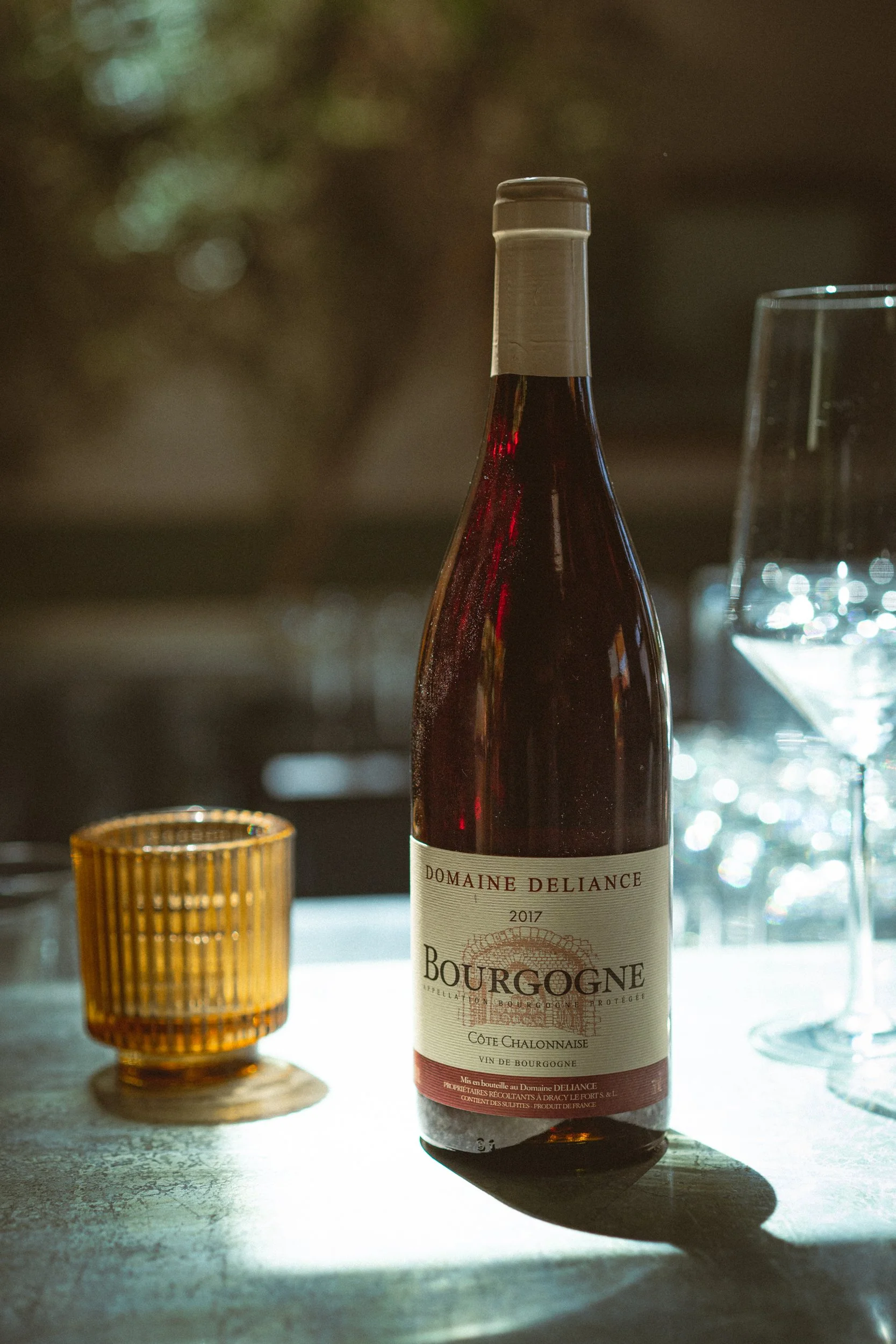 A bottle of Bourgogne wine, vintage 2017, with an amber glass candle holder and a wine glass on a table with sunlight filtering through.