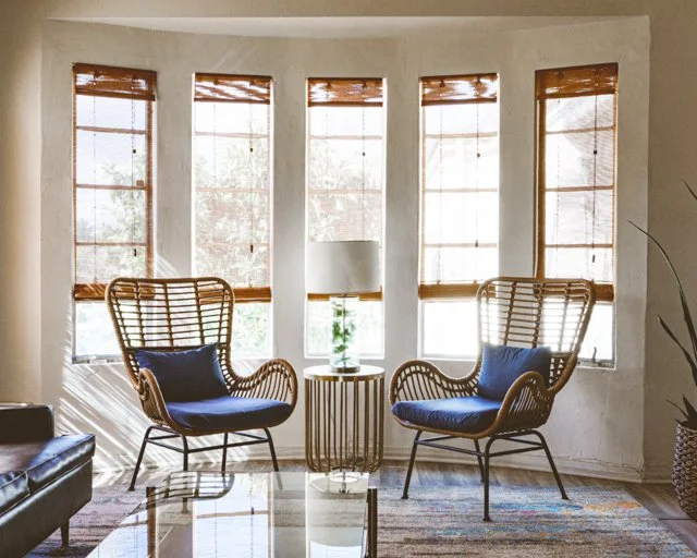 Cozy living room with two wicker chairs with blue cushions, a small round side table with a lamp and plant, and walls with five large windows with bamboo blinds allowing natural light.