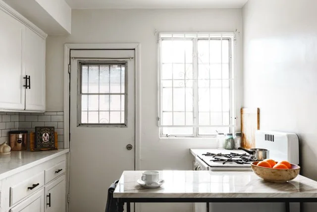 Bright kitchen with white cabinets, a window, a door, a stove, a bowl of oranges, and various kitchen items on the counter.