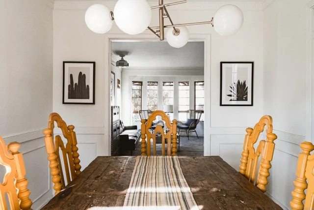 Dining room with a wooden table, yellow chairs, modern chandelier, black and white artwork, and a bright sunlit room visible through a doorway.