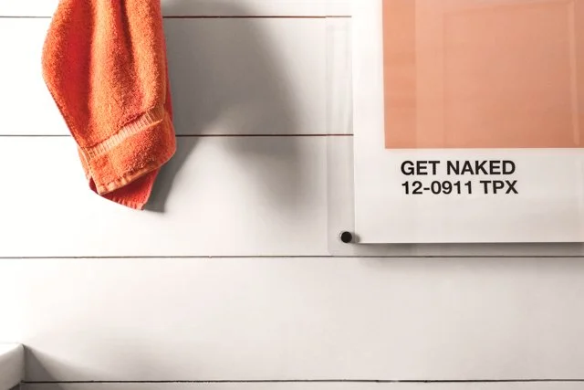 A white wall with horizontal paneling, an orange towel hanging on a hook, and a pink poster with black text reading 'GET NAKED 12-0911 TPX' in the corner.