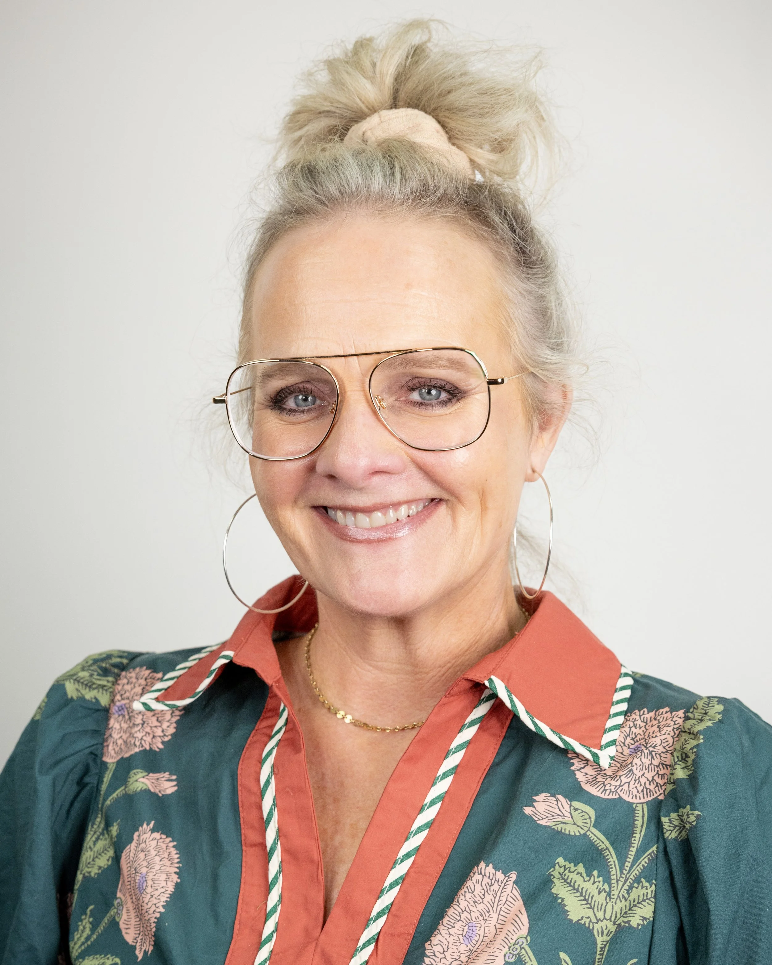 Smiling woman with blonde hair wearing a green blouse and hoop earrings against a plain background.