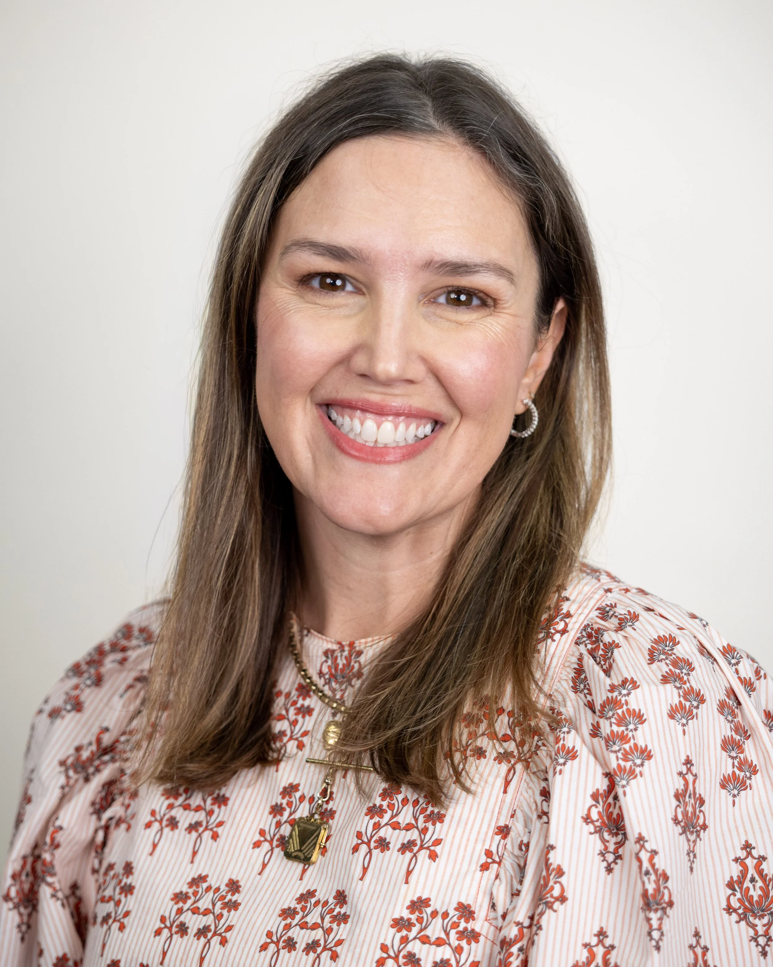 Smiling woman with long brown hair wearing a red floral print dress, posed against a neutral background.