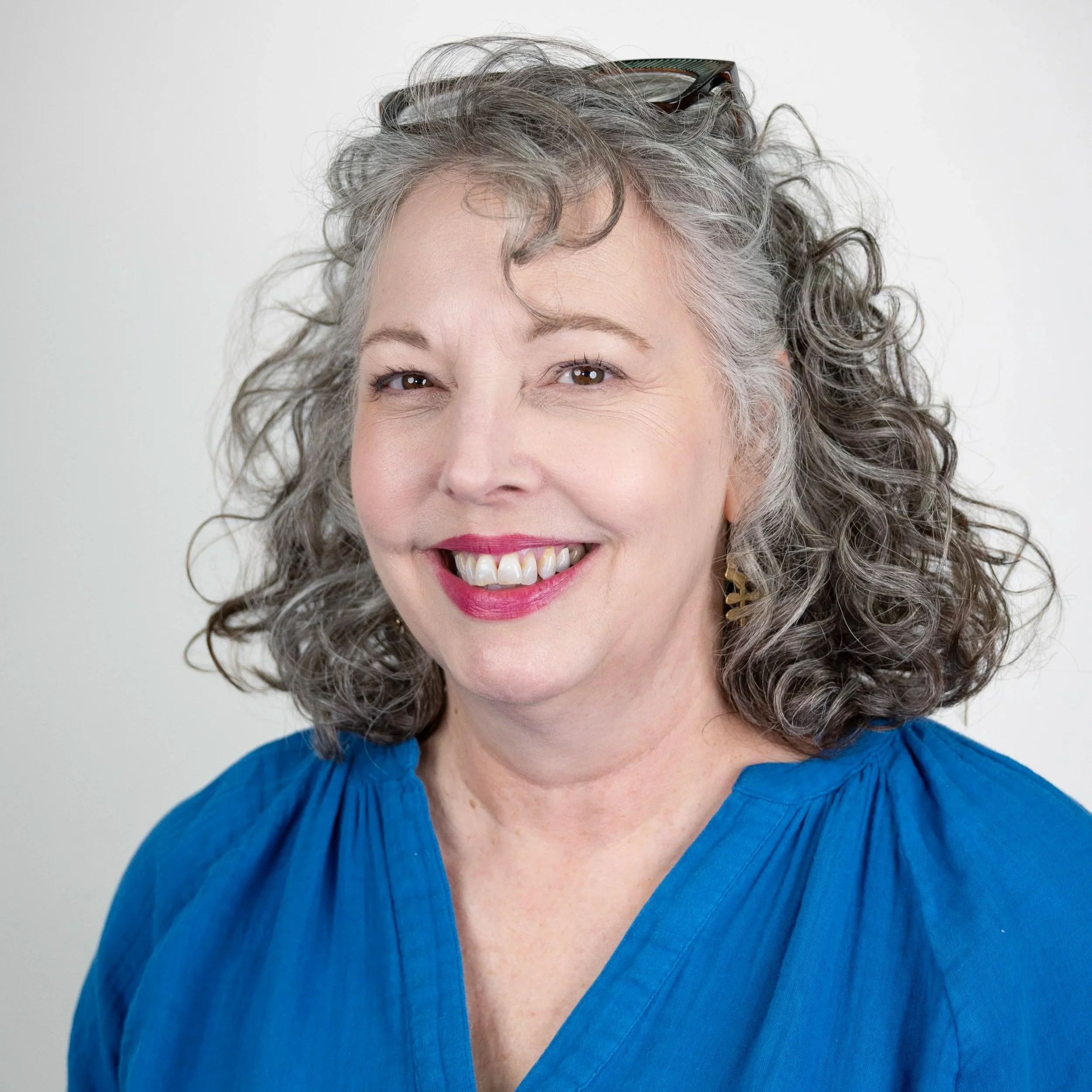Smiling woman with curly gray hair, wearing a white top, against a plain background.