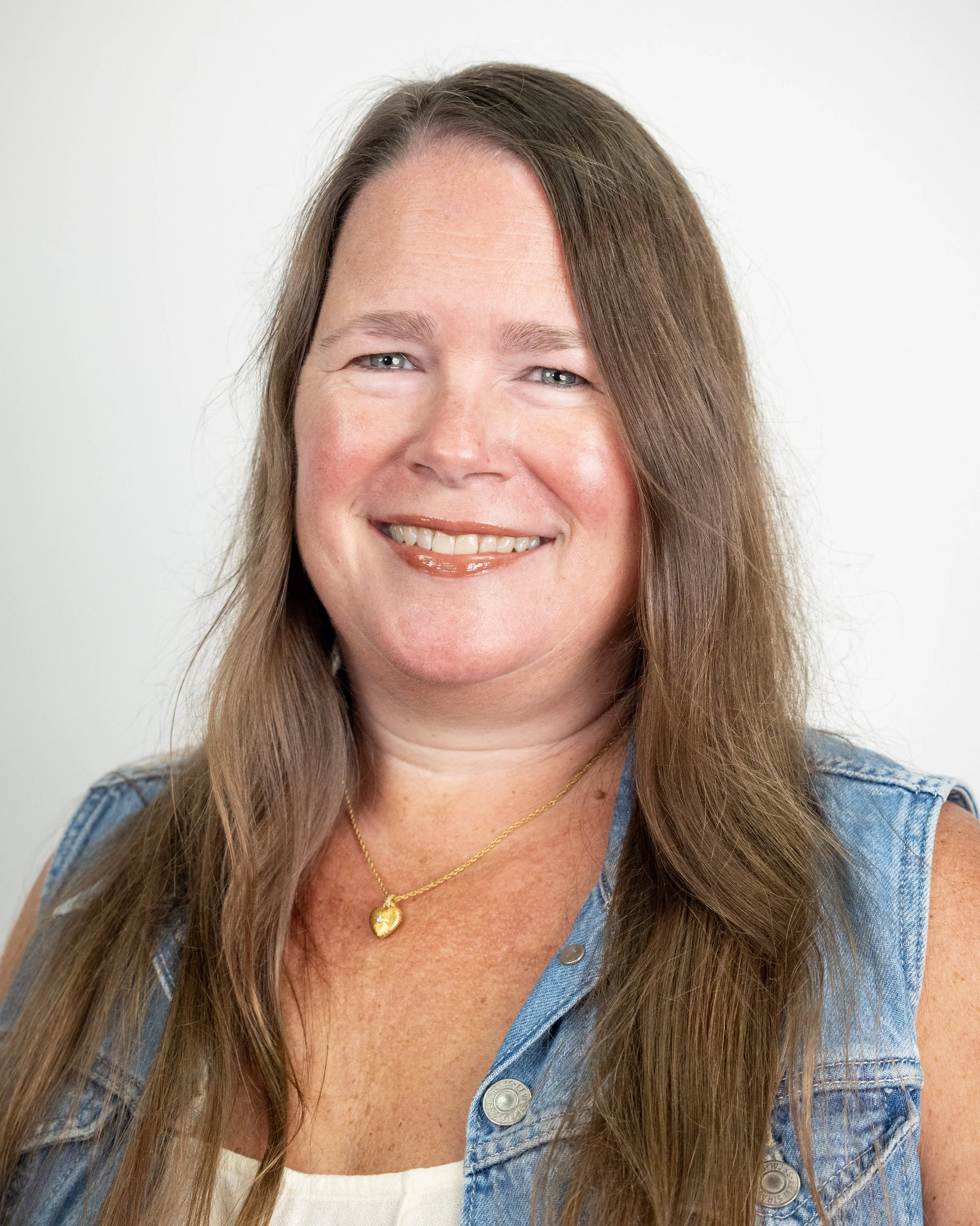 A smiling woman with long brown hair wearing a patterned top and two necklaces.