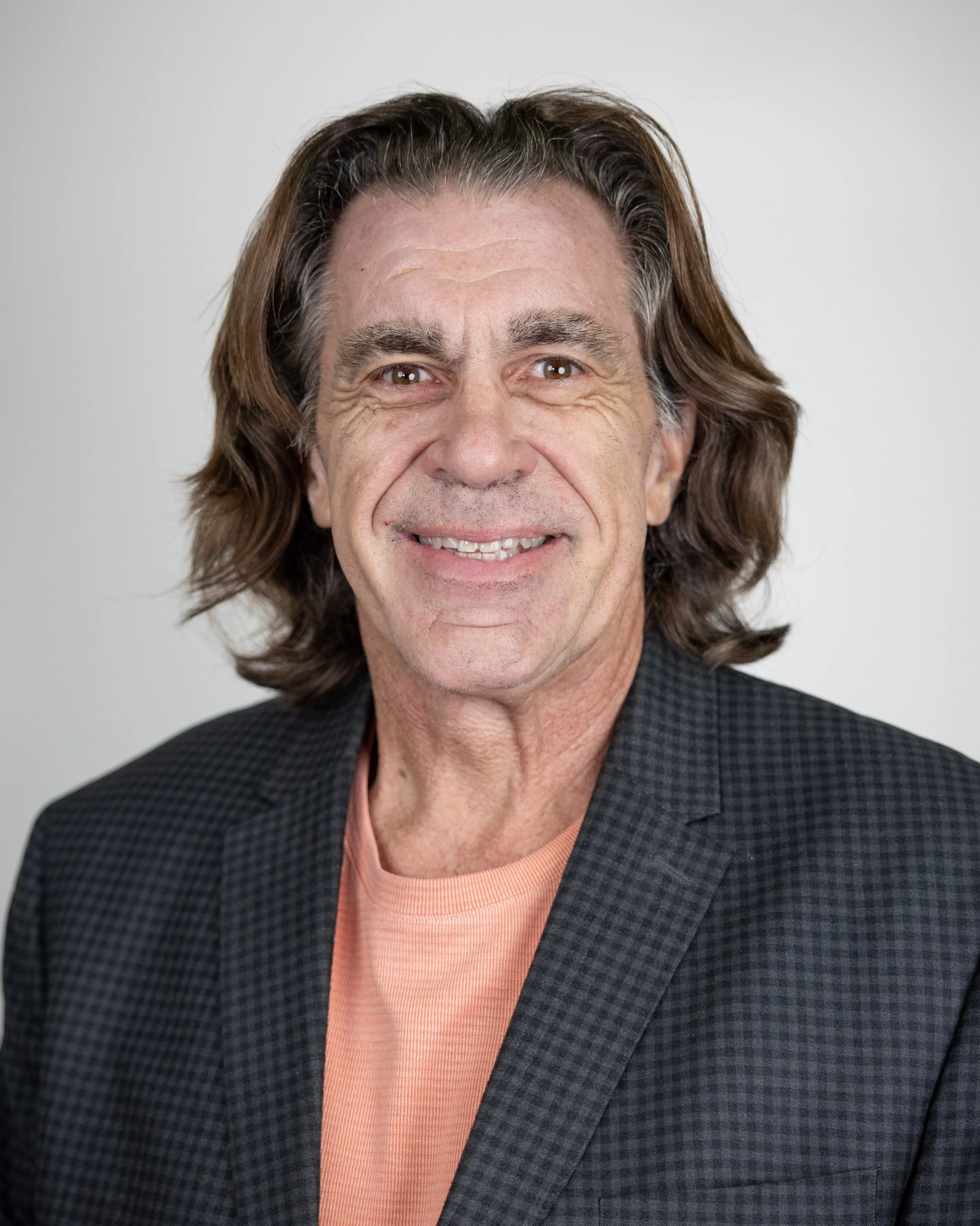 Smiling man with long hair wearing a beige blazer and pink checkered shirt.