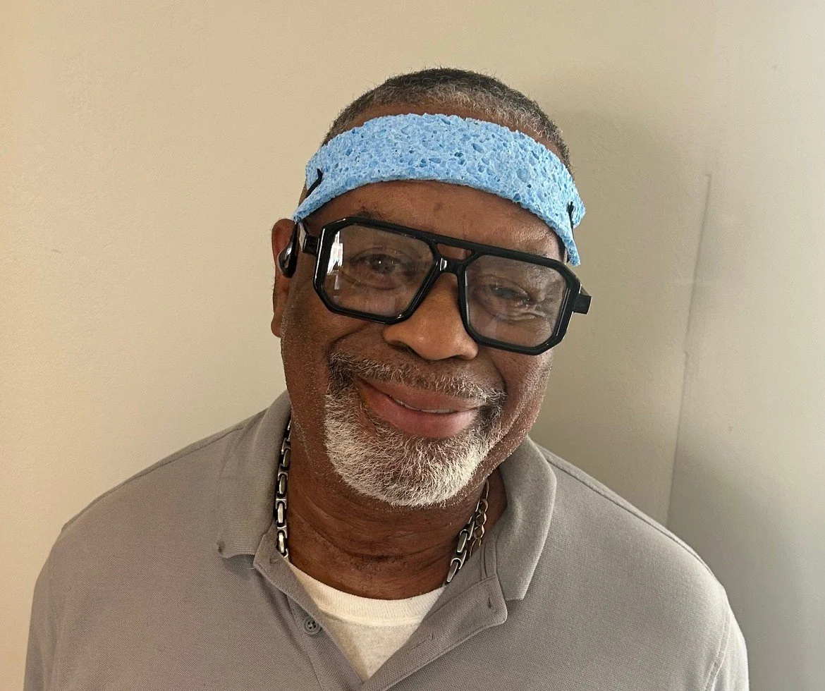 Man wearing a blue headband, glasses, and a blue polo shirt, smiling in front of a textured blue and brown background.