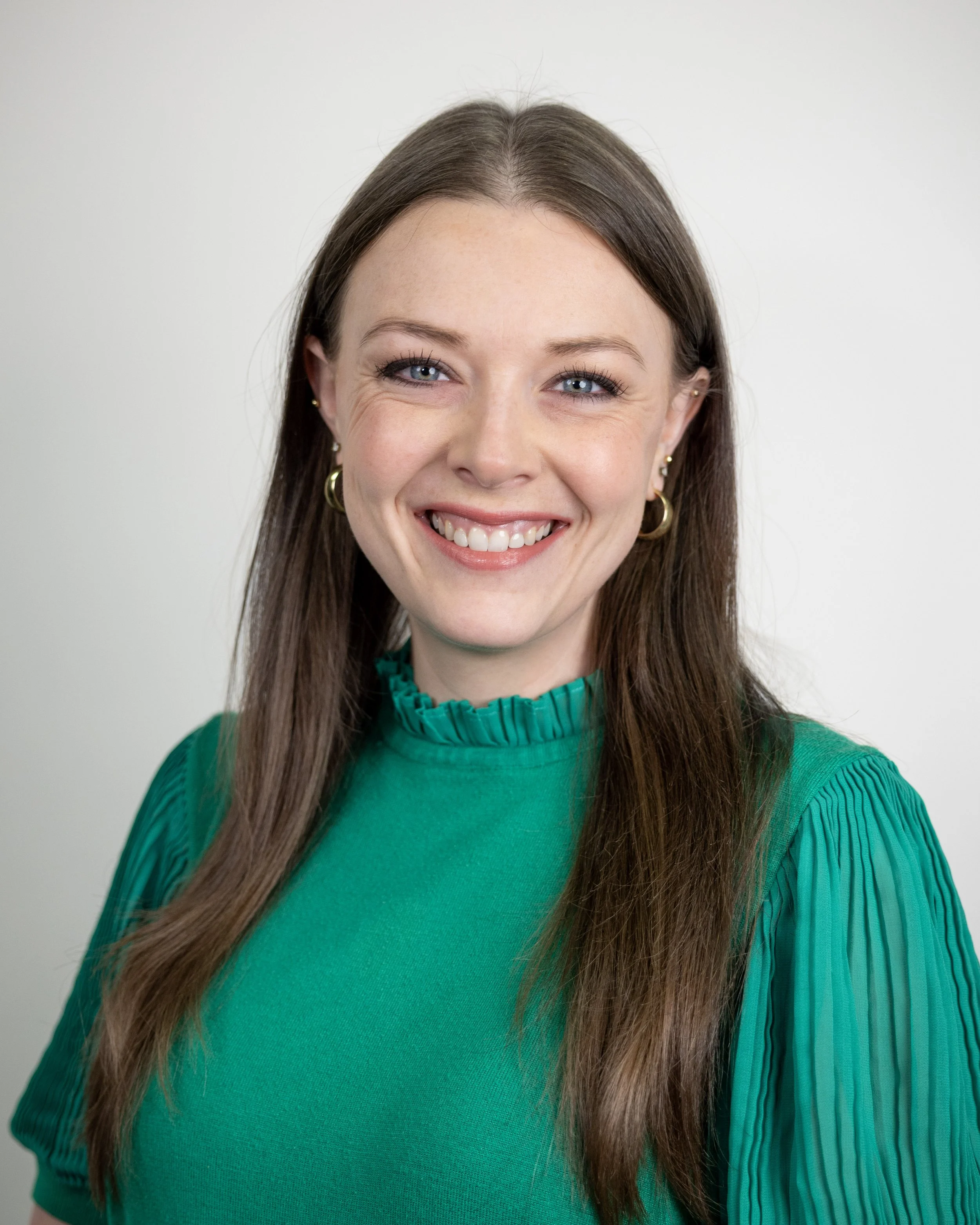 Woman with long brown hair, wearing a white sweater and large brown circular earrings, smiling at the camera.