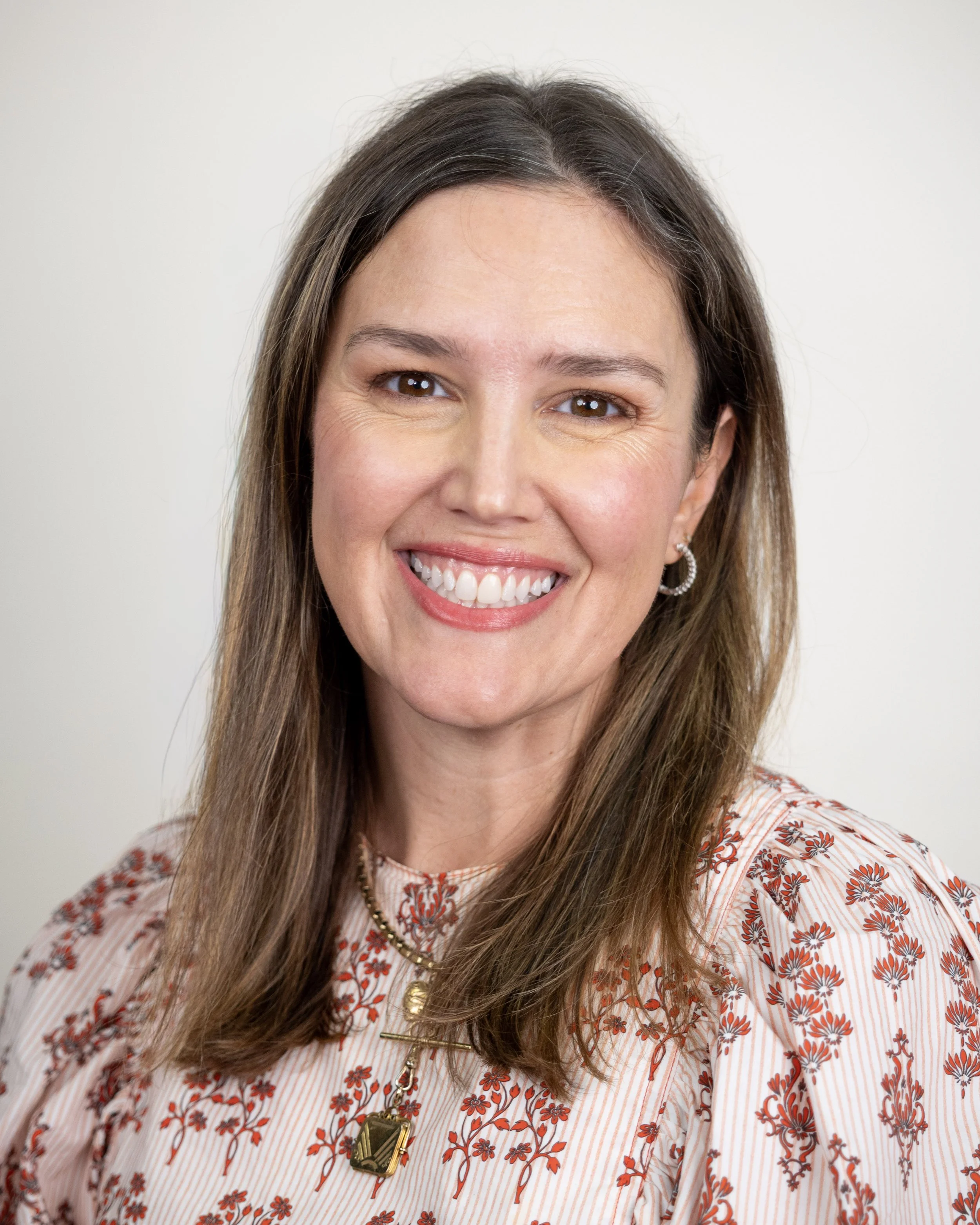 Smiling woman with long brown hair wearing a red floral print dress, posed against a neutral background.