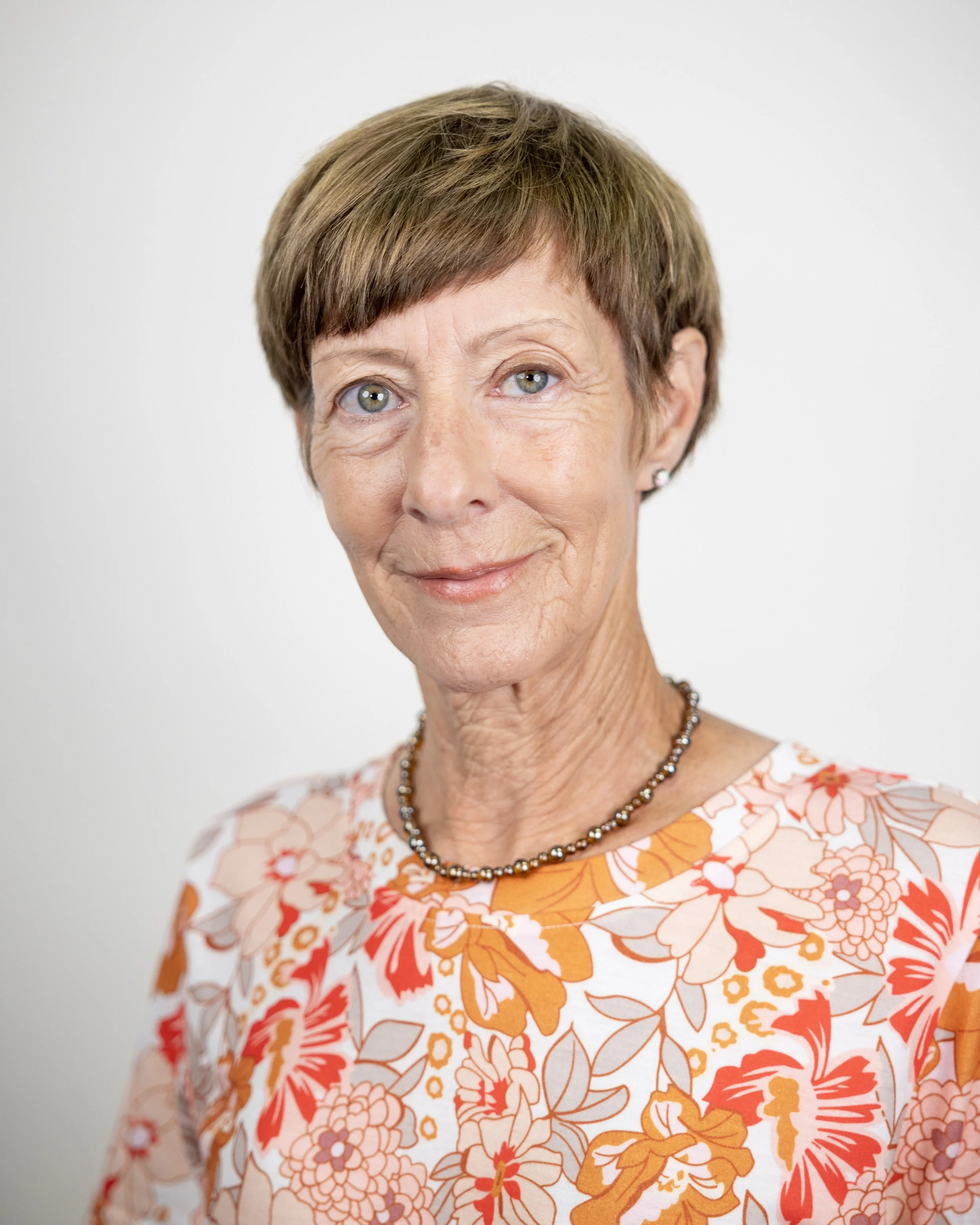 A person with short brown hair wearing a green, patterned shirt, smiling against a plain background.