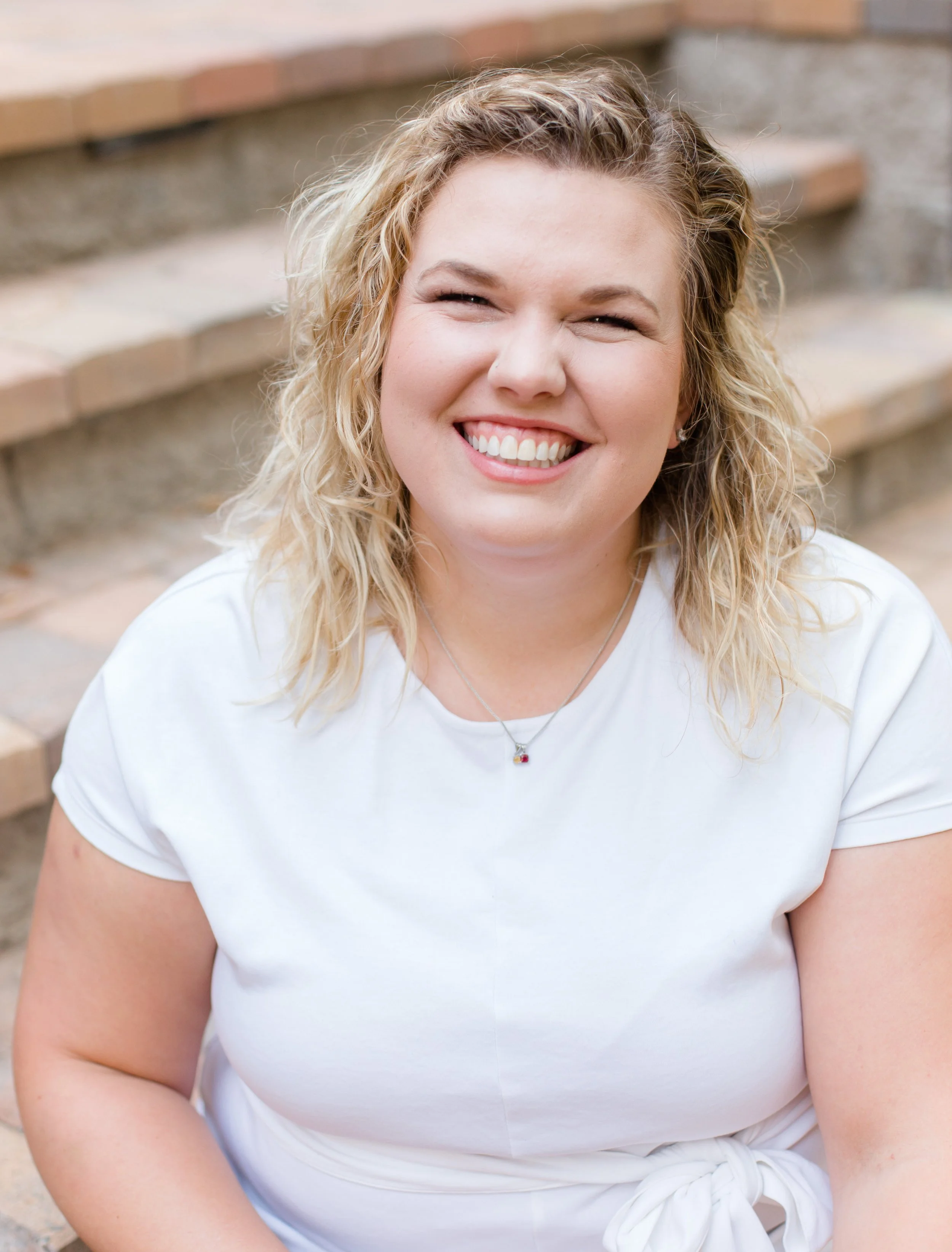A smiling woman with curly blonde hair sitting on outdoor brick stairs, wearing a white t-shirt and a small necklace.