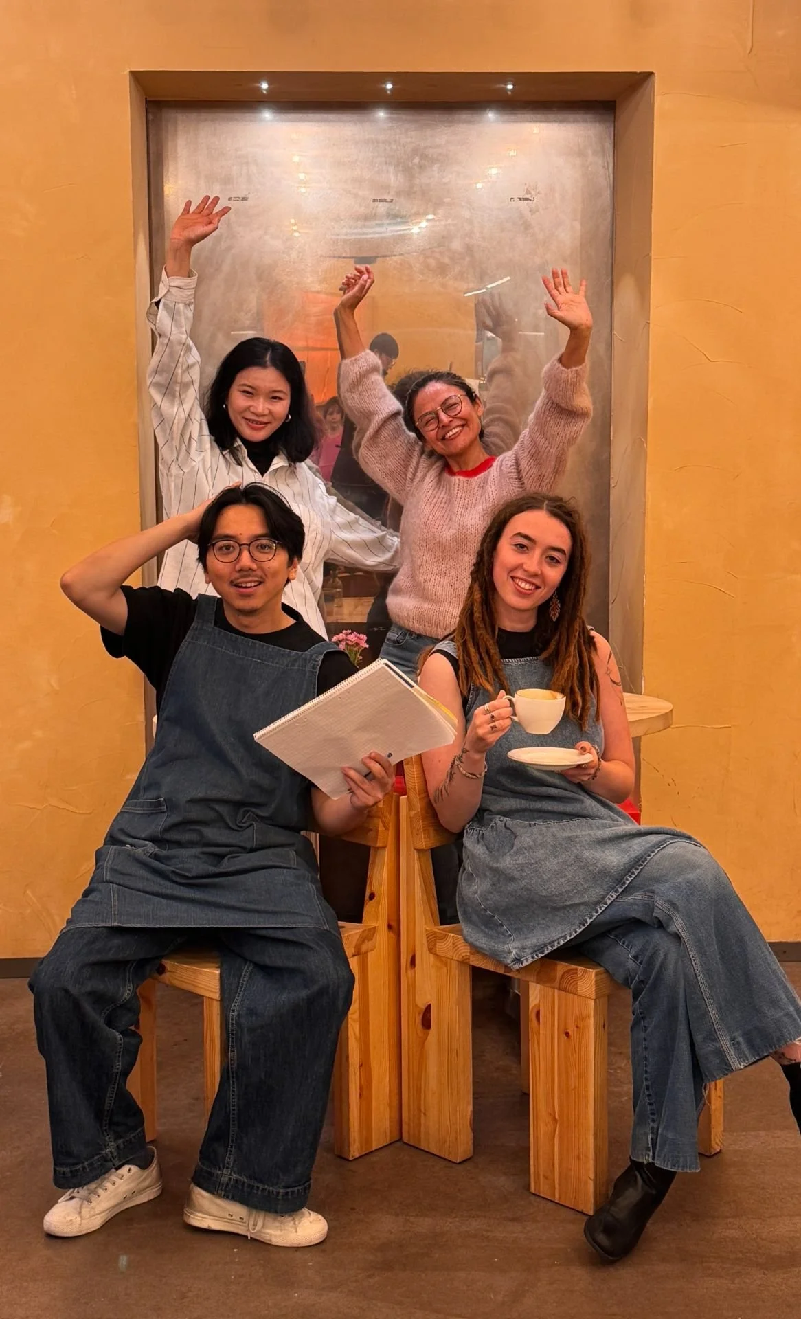 Four friends smiling and posing for a photo in a cozy cafe. Two are sitting on wooden benches, one holding a notebook, the other holding a cup of tea, while two stand behind them with their hands raised cheerfully.