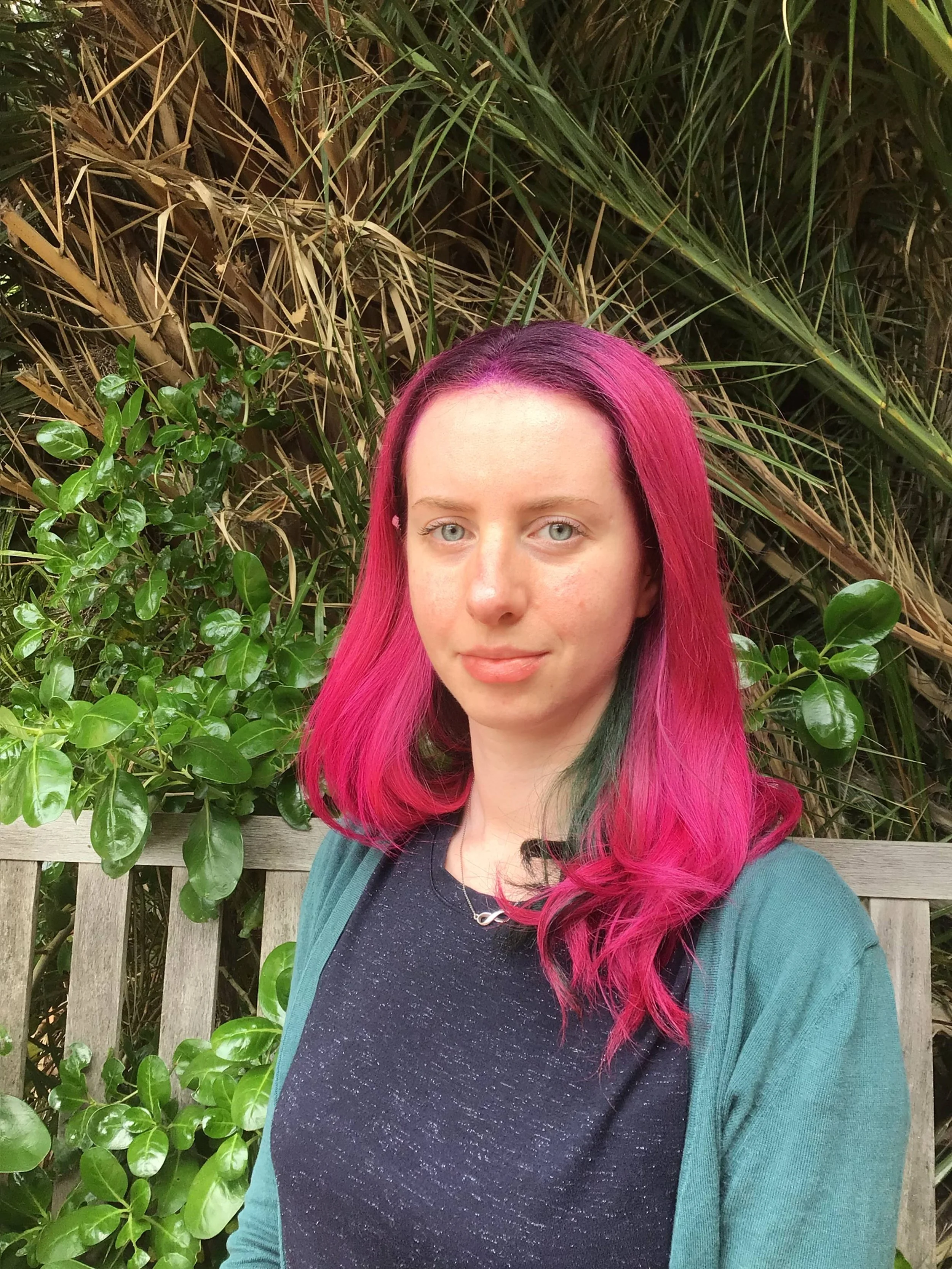 A young woman with pink hair and light eyes standing outdoors in front of dense green foliage and a wooden fence.