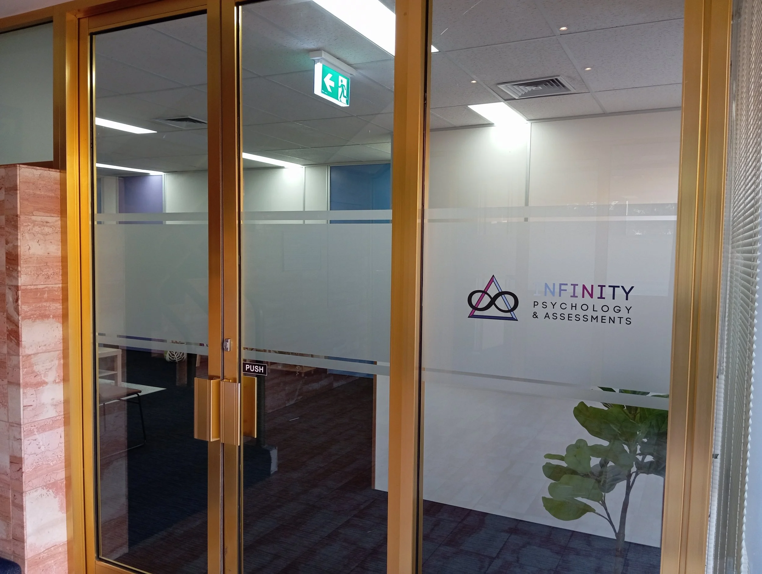 Glass door with yellow frame leading to an office space with a sign for 'Infinity Psychology & Assessments' visible through the glass door, and a green exit sign with a running figure above.