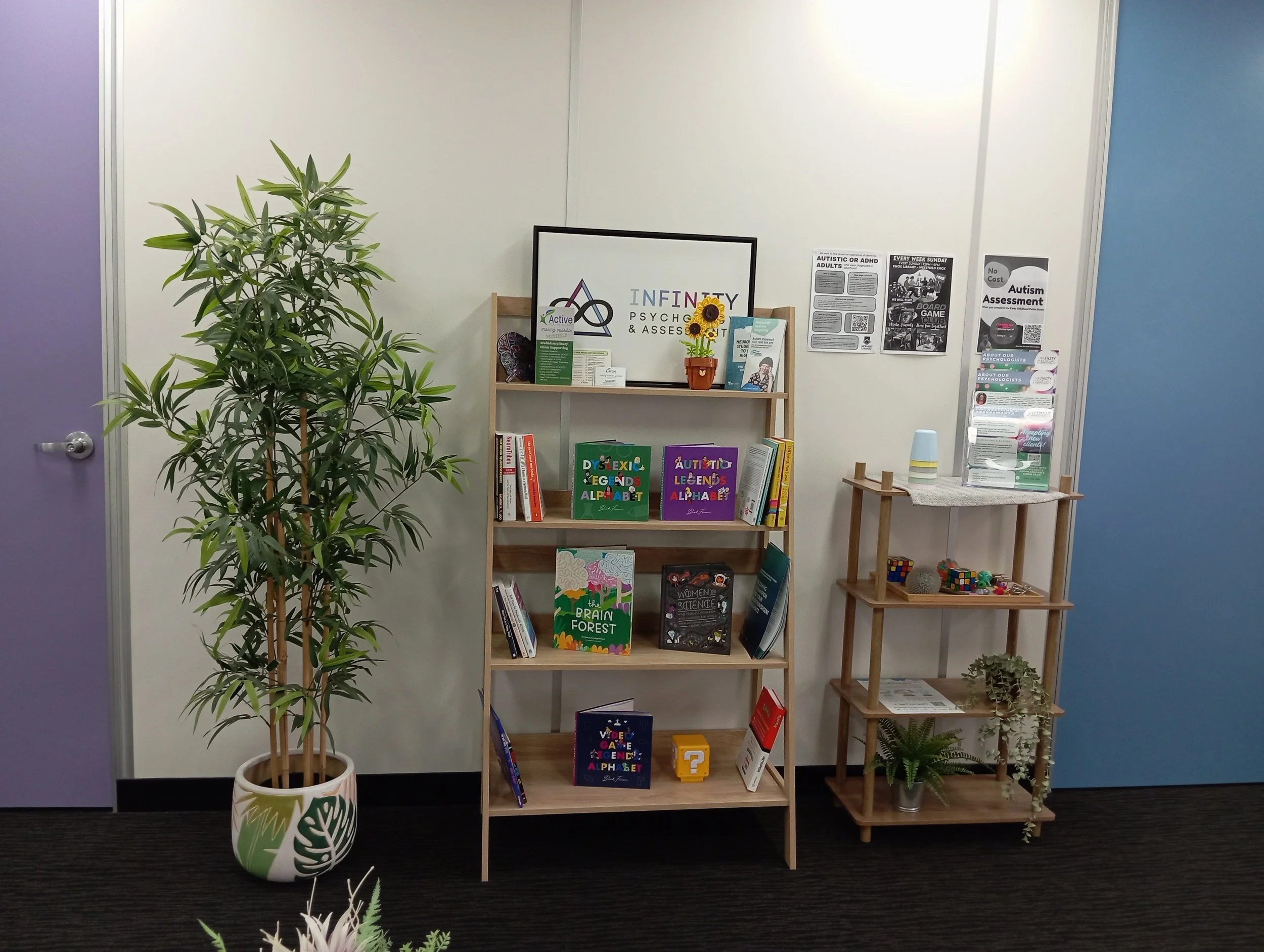 A bookstore or library display with a large potted plant on the left, a white wall with various posters on the right, a bookshelf filled with colorful books about autism and dyslexia, and two smaller shelves holding additional books and decorative it
