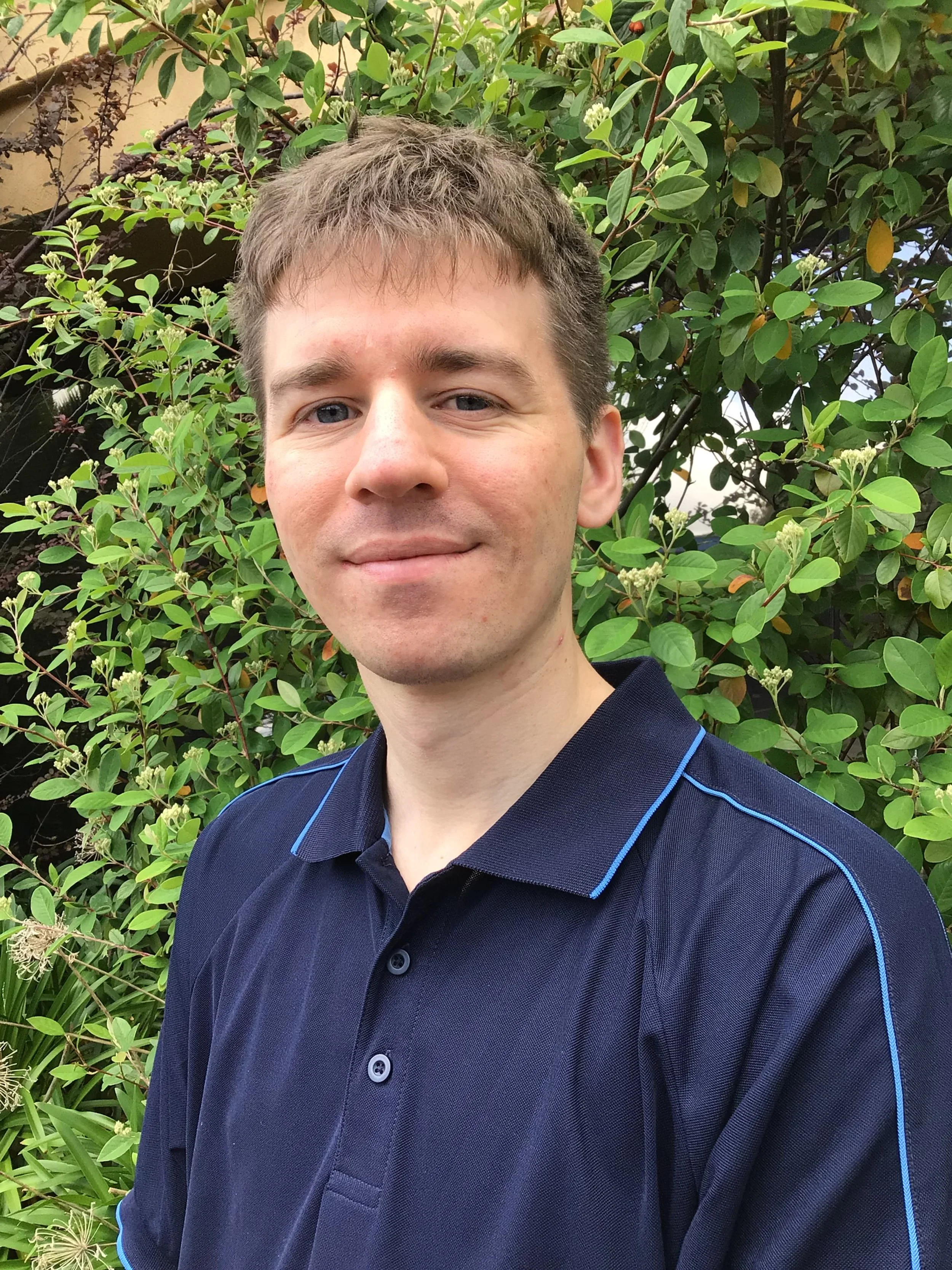 A young man with short light brown hair and blue eyes taking a selfie in front of green leafy bushes in a garden.