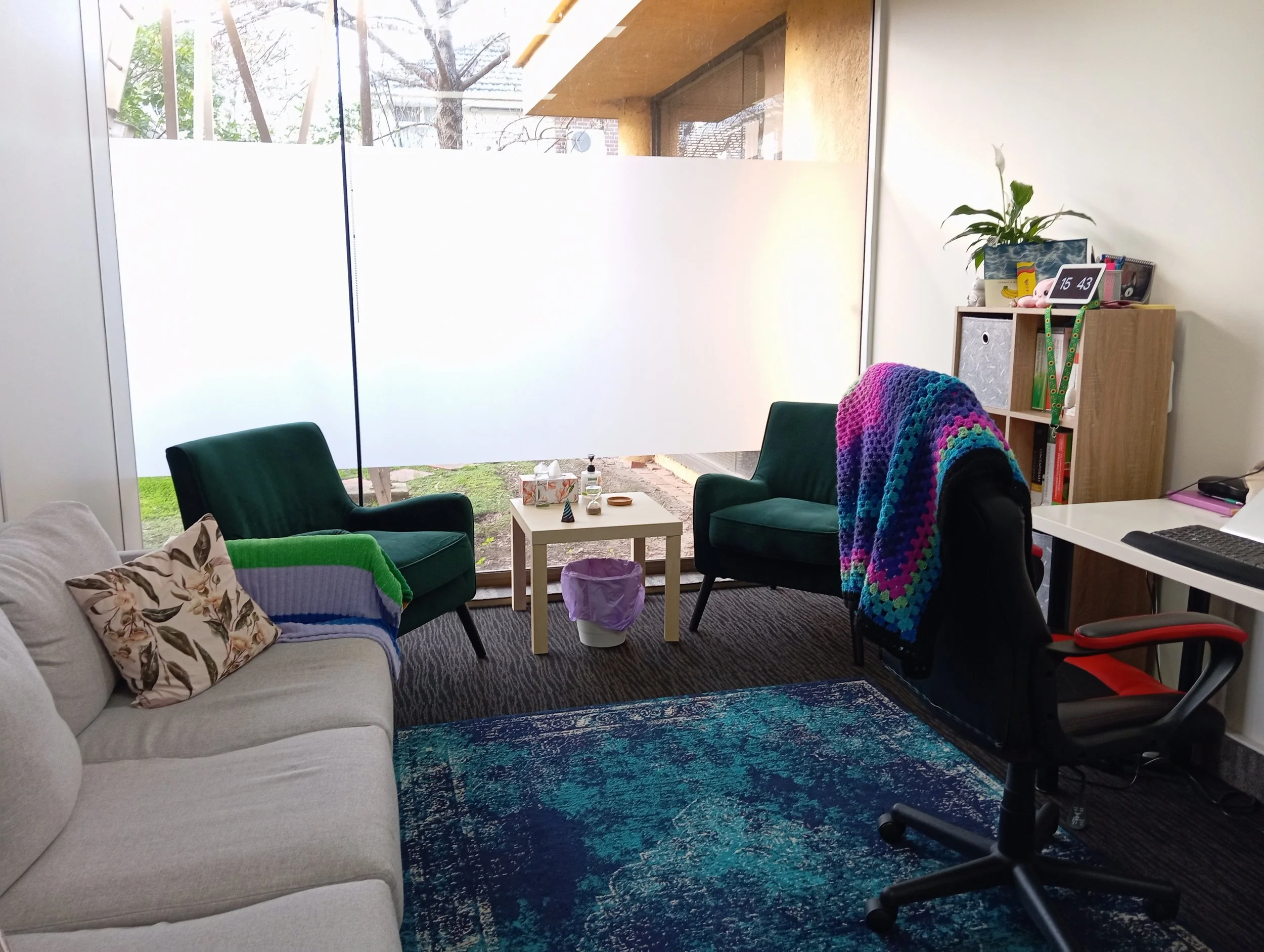 A cozy room with a large window looking out to a backyard with trees. The room has a white sofa with floral and green pillows, two dark green armchairs, a small white coffee table with decorative items, a bookshelf with books and a plant, and a fuzzy