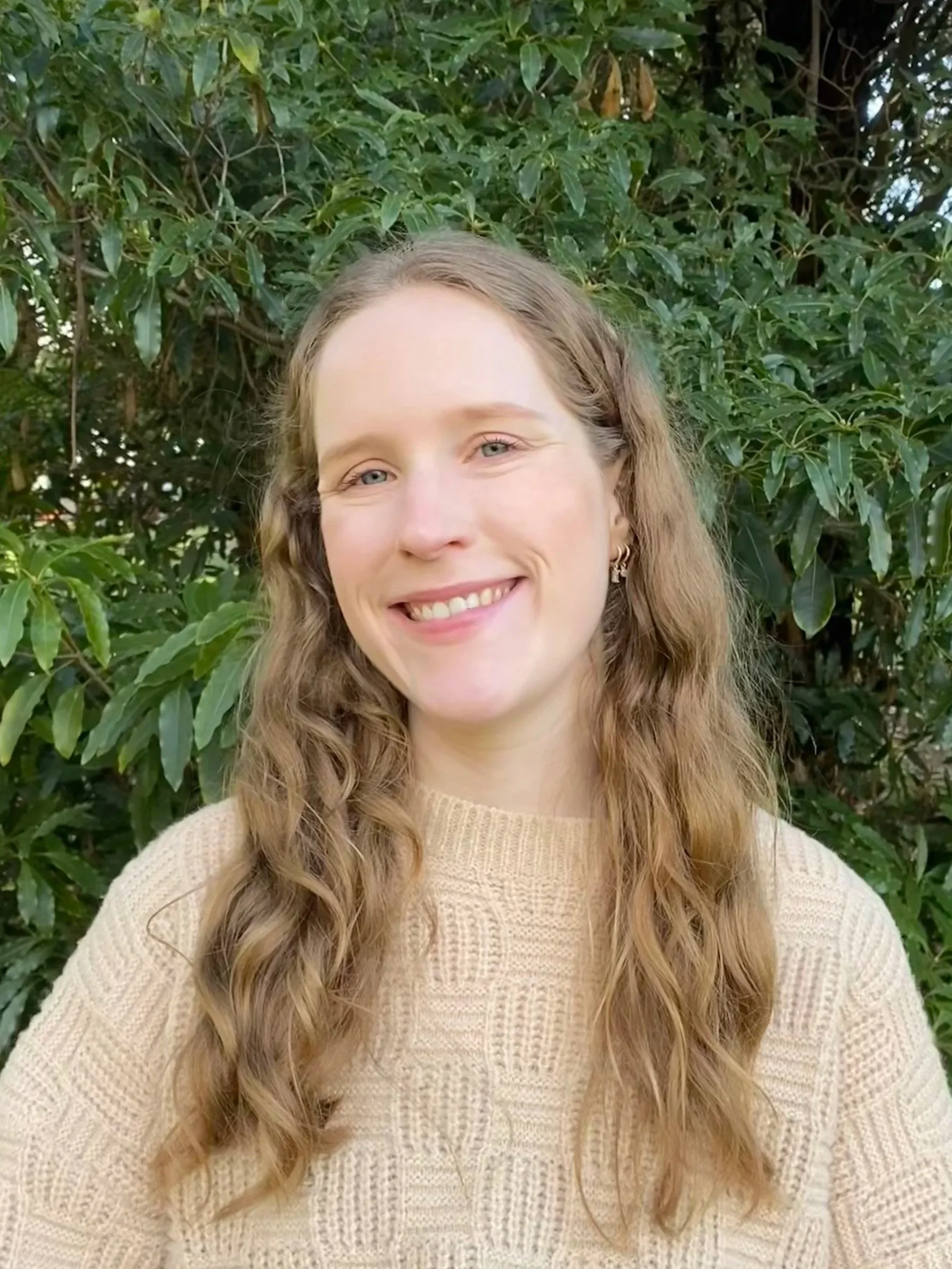 A woman with long wavy blond hair smiling outdoors in front of green foliage, wearing a cream-colored cable knit sweater.