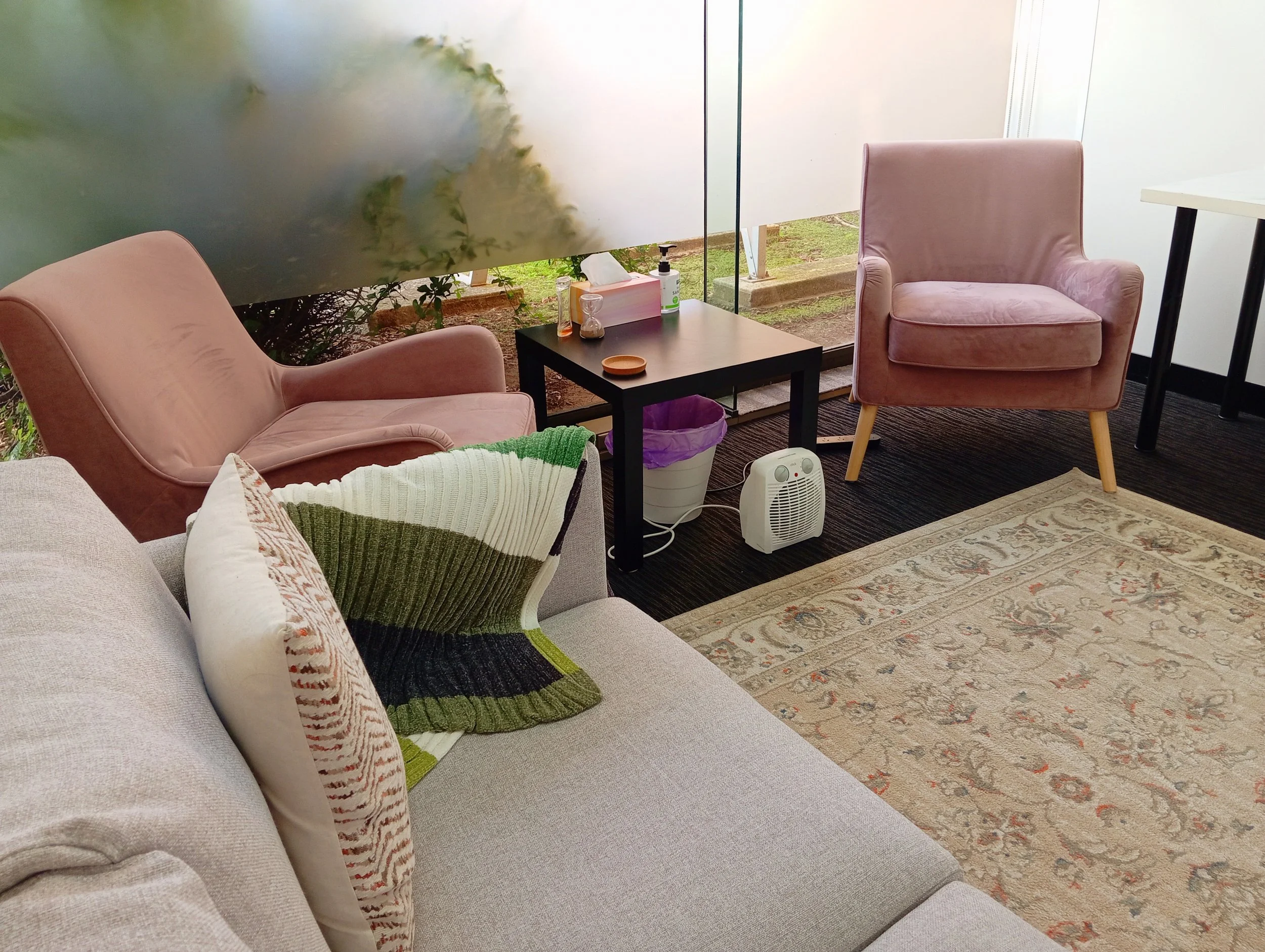 A cozy seating area with pink armchairs, a black coffee table, and a white sofa with decorative pillows, near a large glass window with a view of the outdoors.