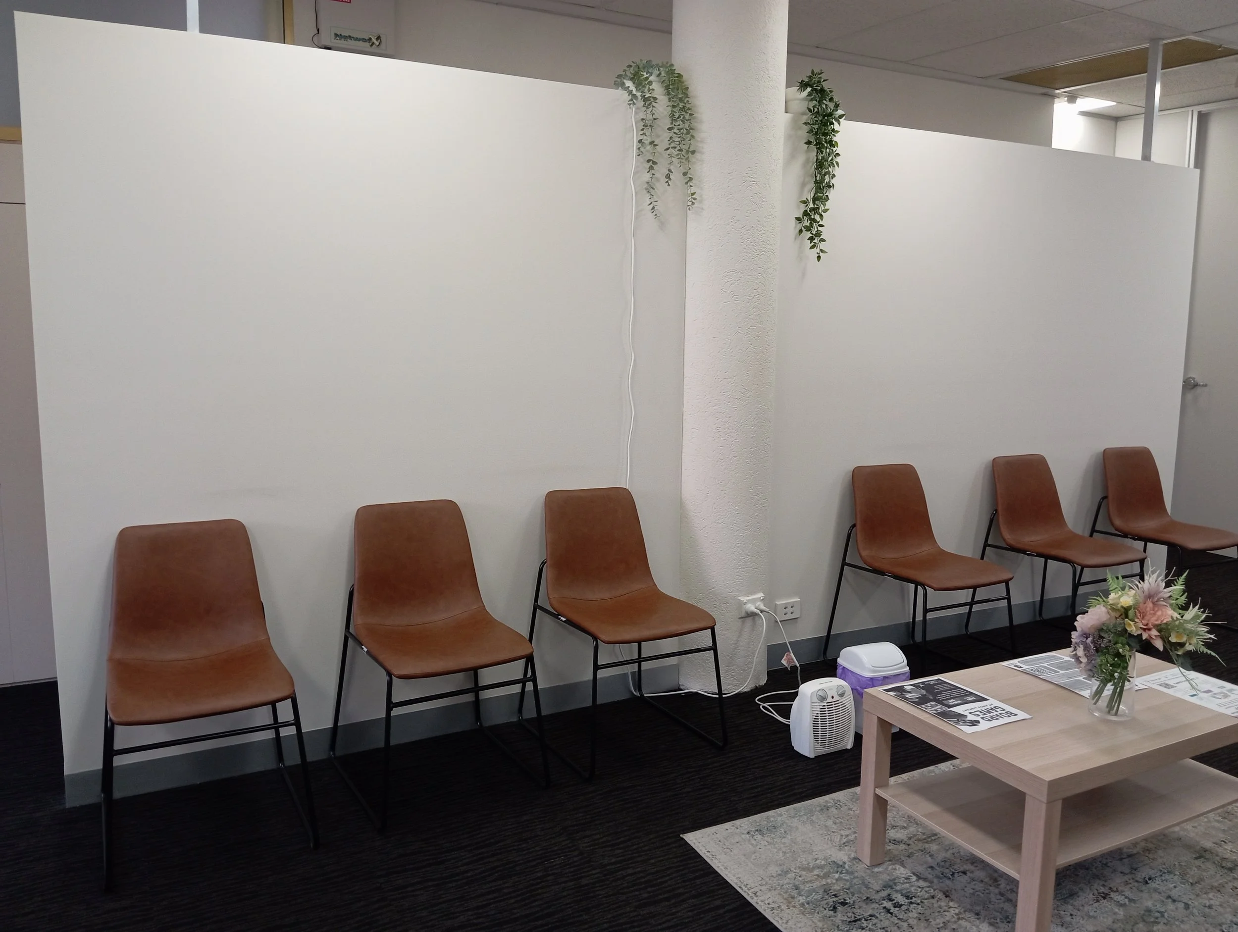 A row of brown chairs against a white partition wall with some hanging plants, a table with flowers, and some electronic devices in an indoor setting, possibly an office or waiting area.