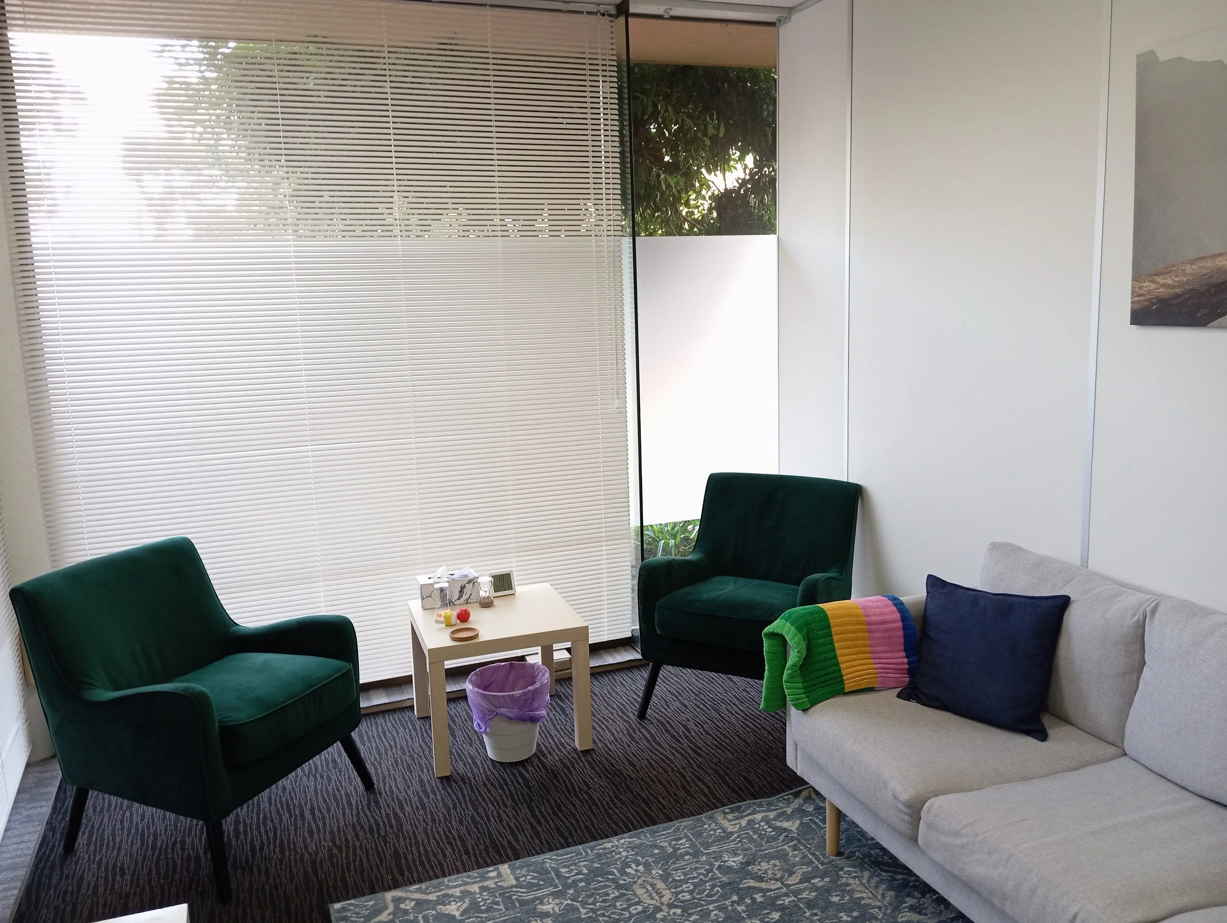 A cozy room with white walls and large windows with blinds. There are four chairs: two green velvet armchairs, a dark armchair with a multicolored blanket, and a gray sofa. A small white table with toys and a digital clock is in the center. A white t