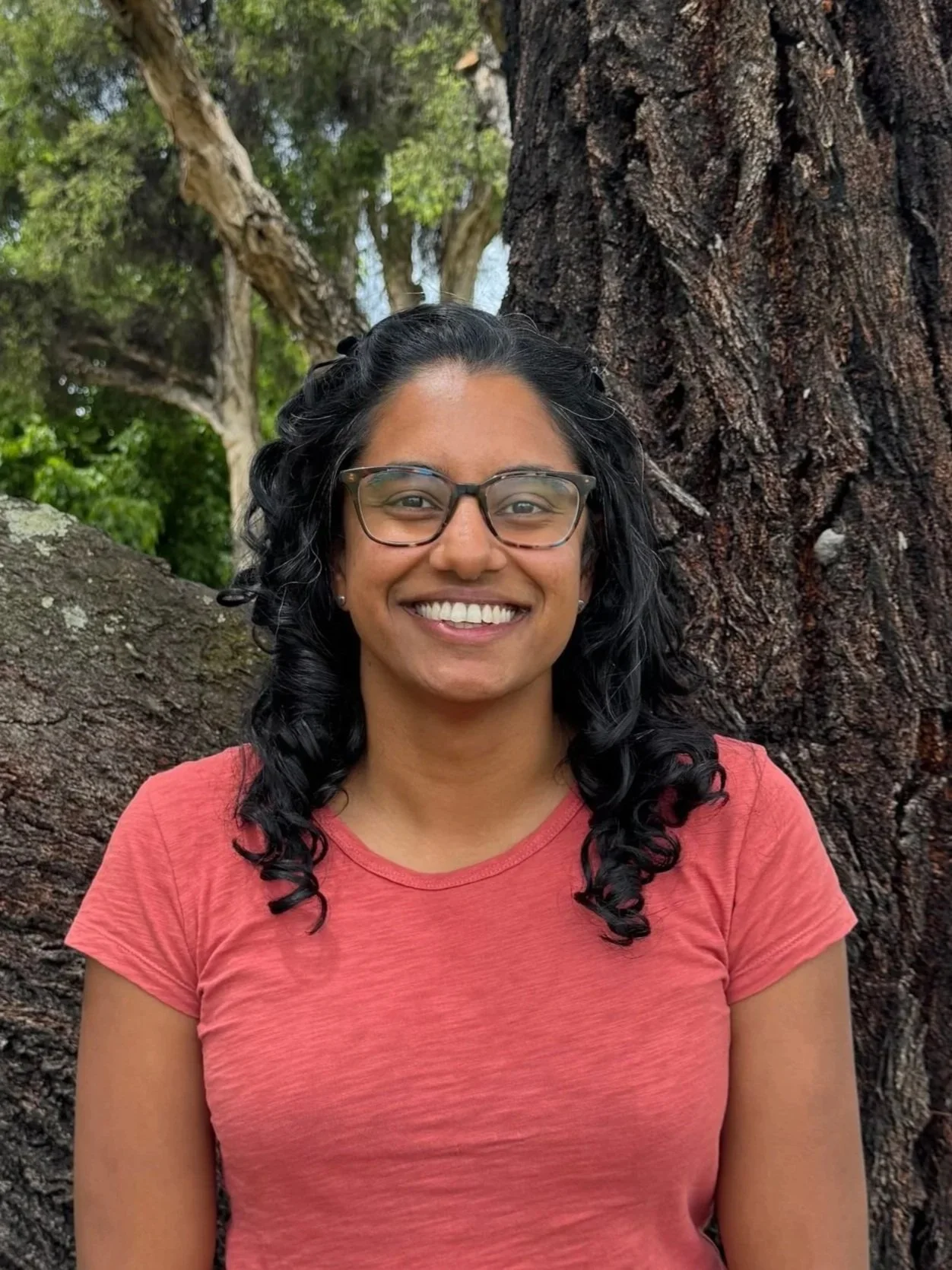 A woman with glasses and curly black hair smiling in front of a tree outdoors.