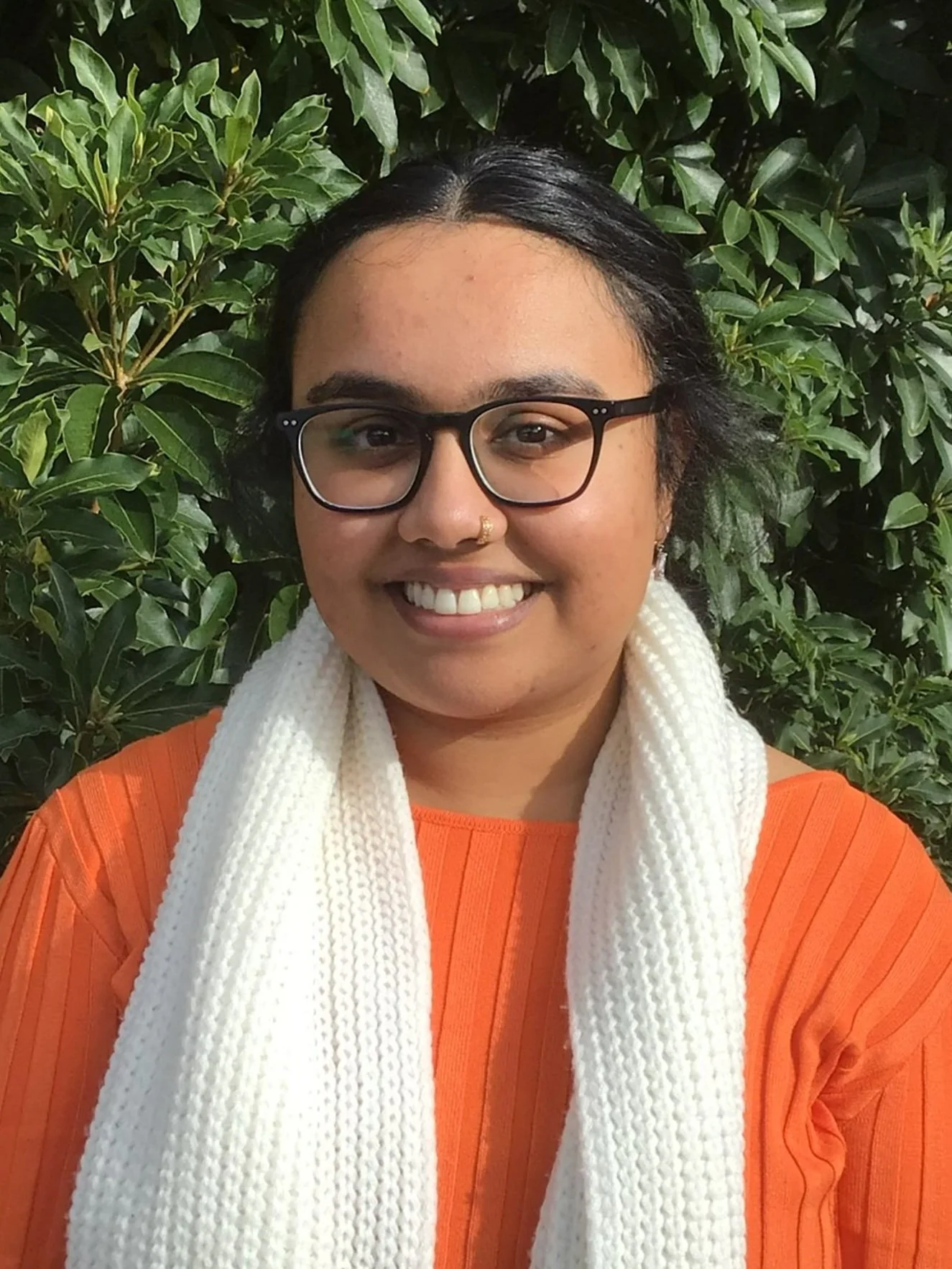A woman with short black hair, glasses, and a wide smile, wearing an orange top and a white scarf, standing in front of green foliage.