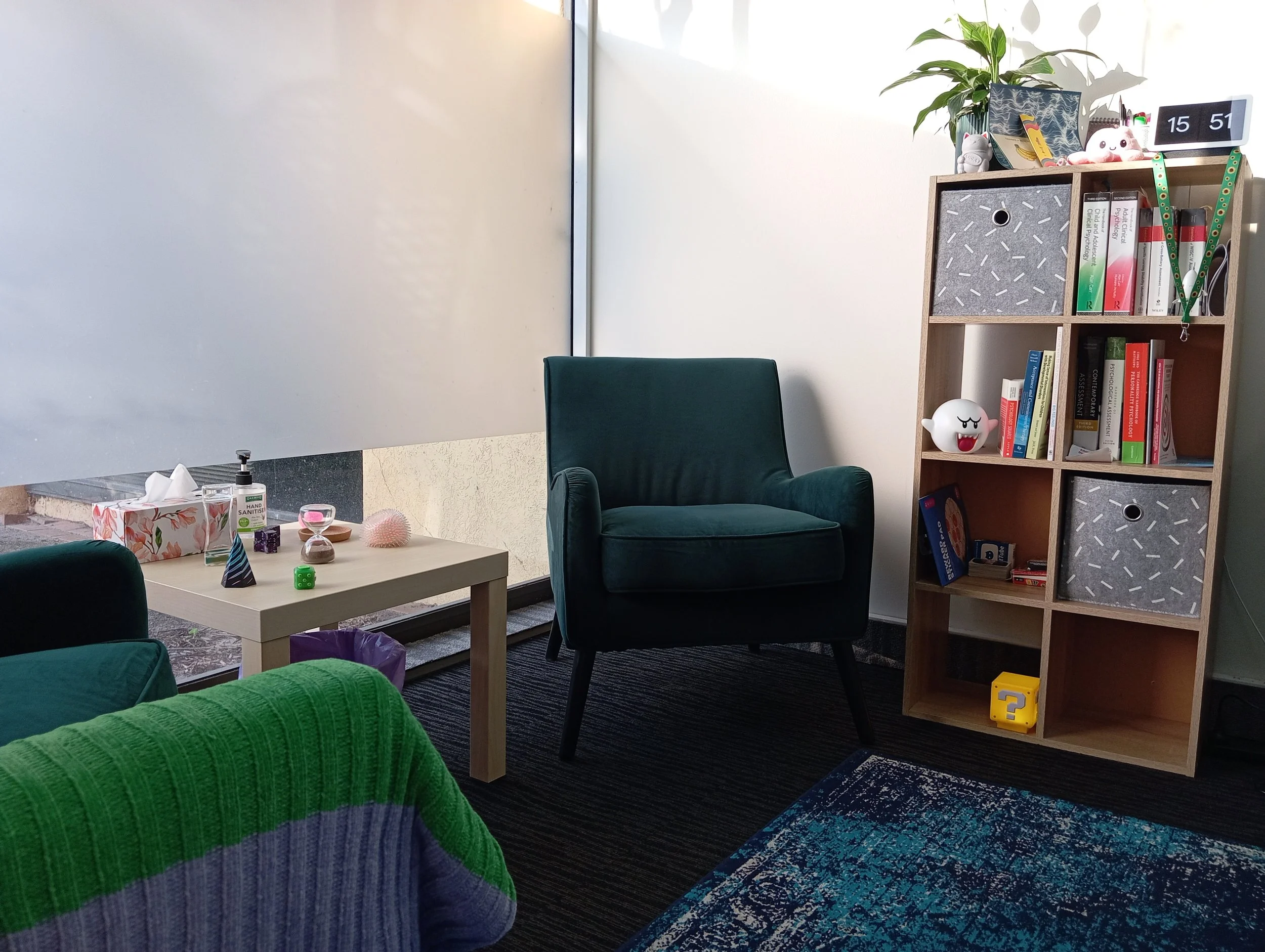 An office with a dark green armchair, a small table with decorations and a tissue box, a wooden bookcase with books and toys, and a white wall with a large whiteboard. Sunlight is coming through the window.