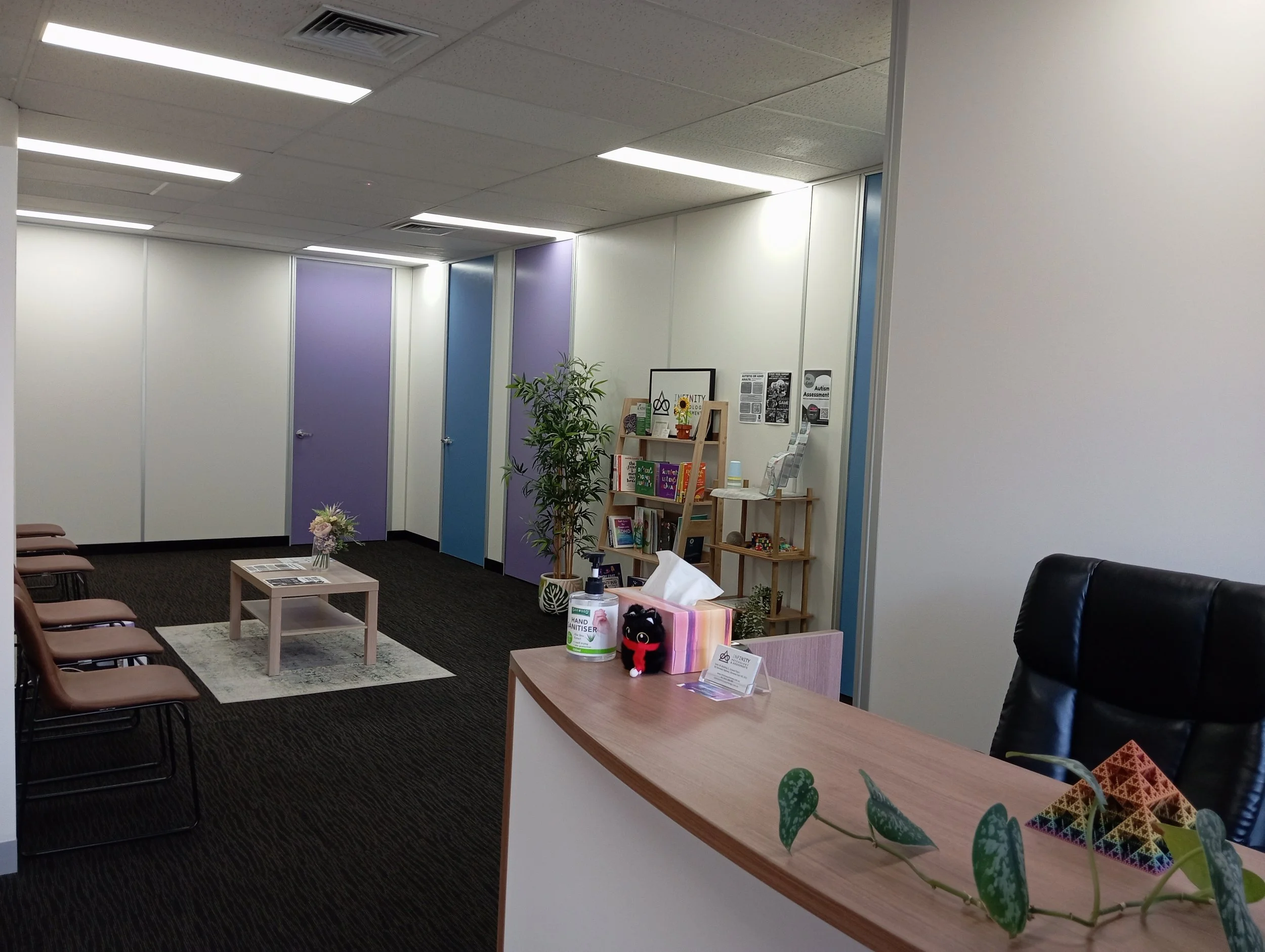 Interior of a waiting room or office area with chairs along the wall, small tables with decorative items, a bookshelf filled with books and magazines, plants, and a white wall with posters. The ceiling has fluorescent lights and air vents.