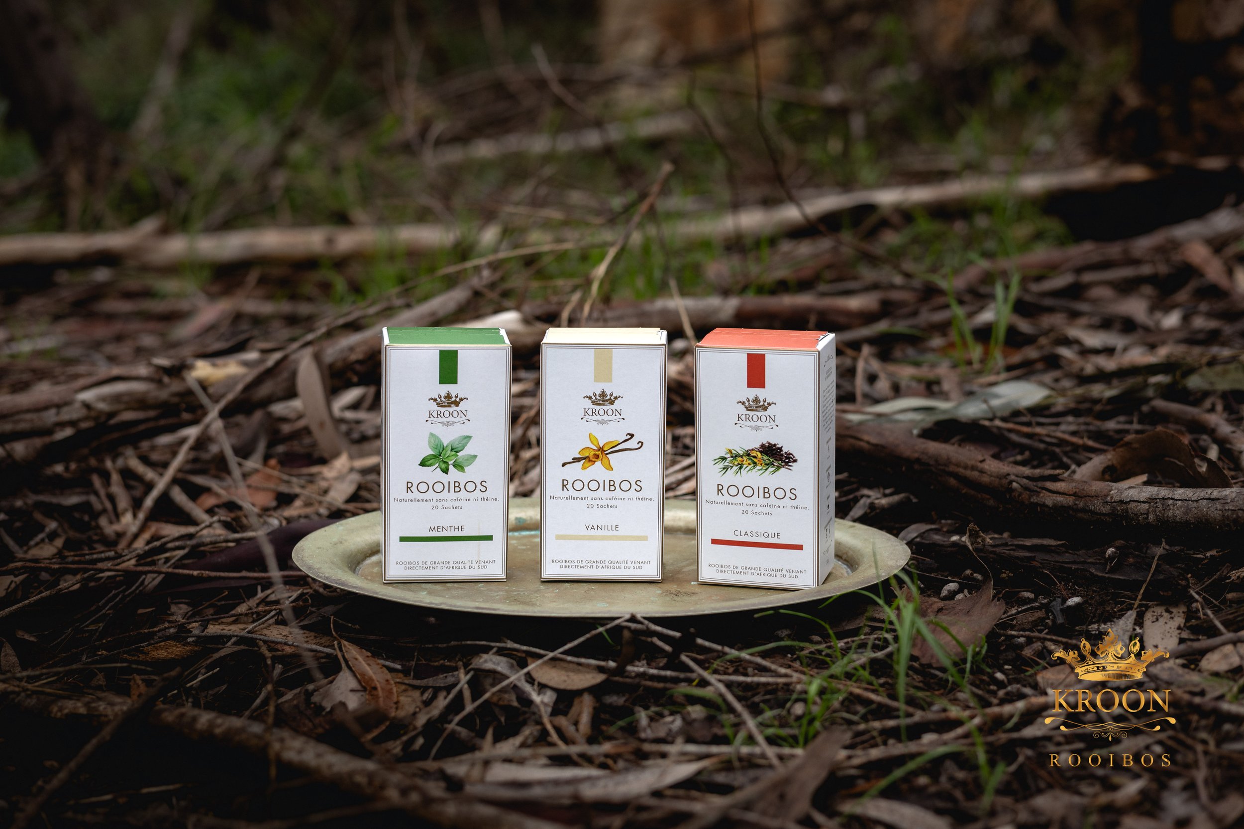 Trois paquets de rooibos sur une assiette en plein air, entourés de feuilles et de branches, portant des étiquettes menthe, vanille, et classique.