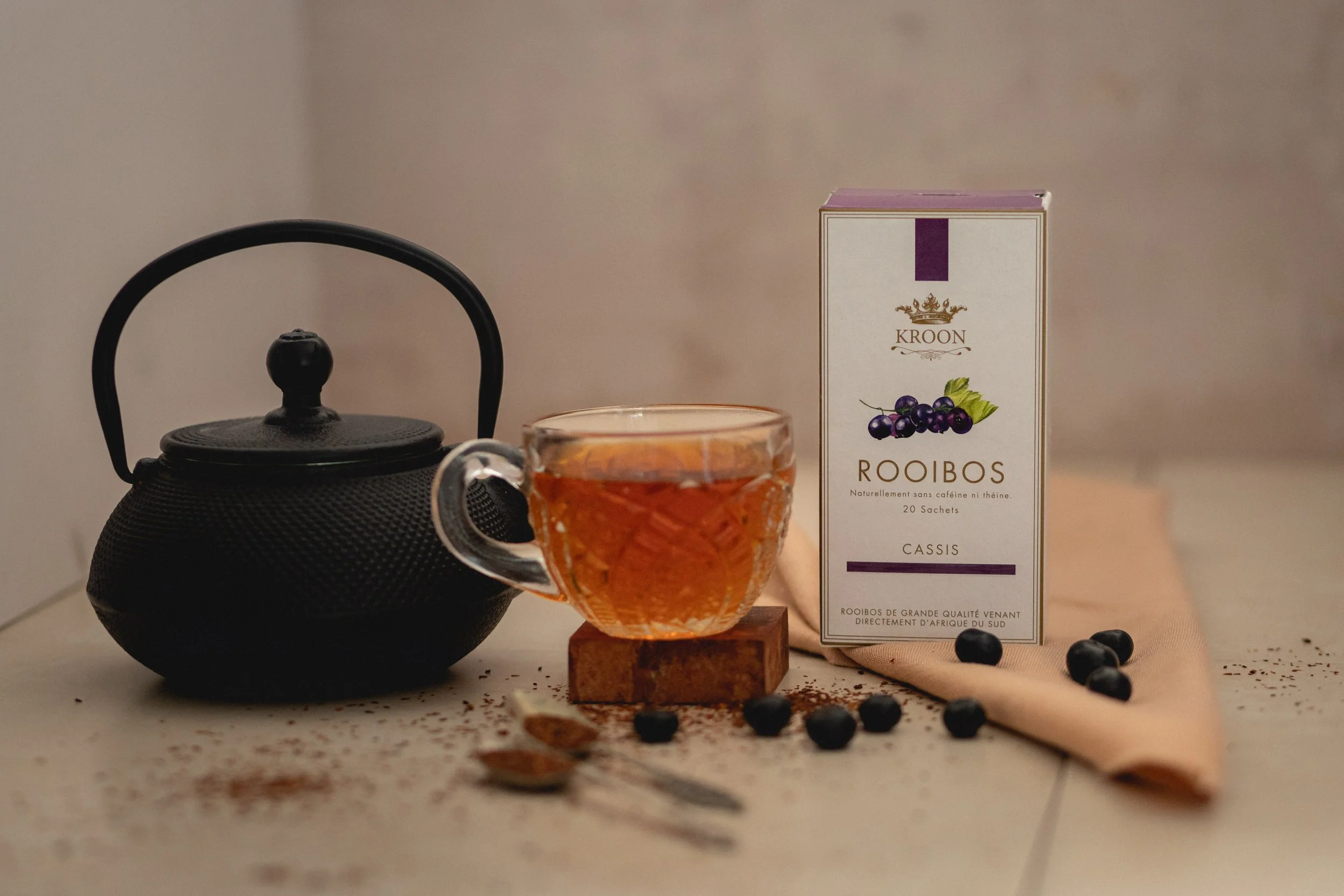 Une théière noire, une tasse en verre avec du thé, une boîte de rooibos cassis et quelques baies de cassis avec une cuillère sur une nappe beige.
