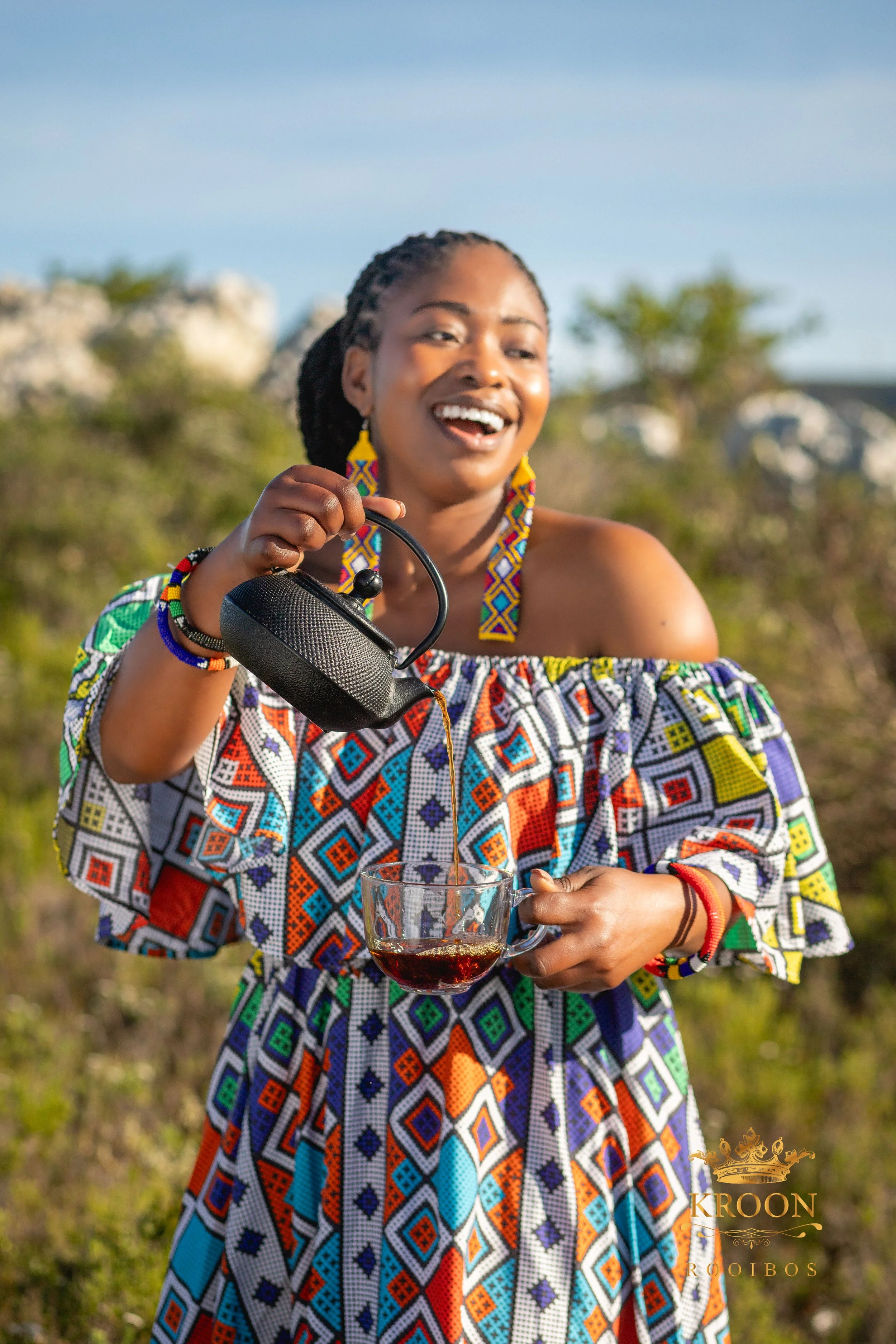 Une femme souriante portant des vêtements traditionnels colorés verse du vin dans un verre lors d'une journée en plein air.