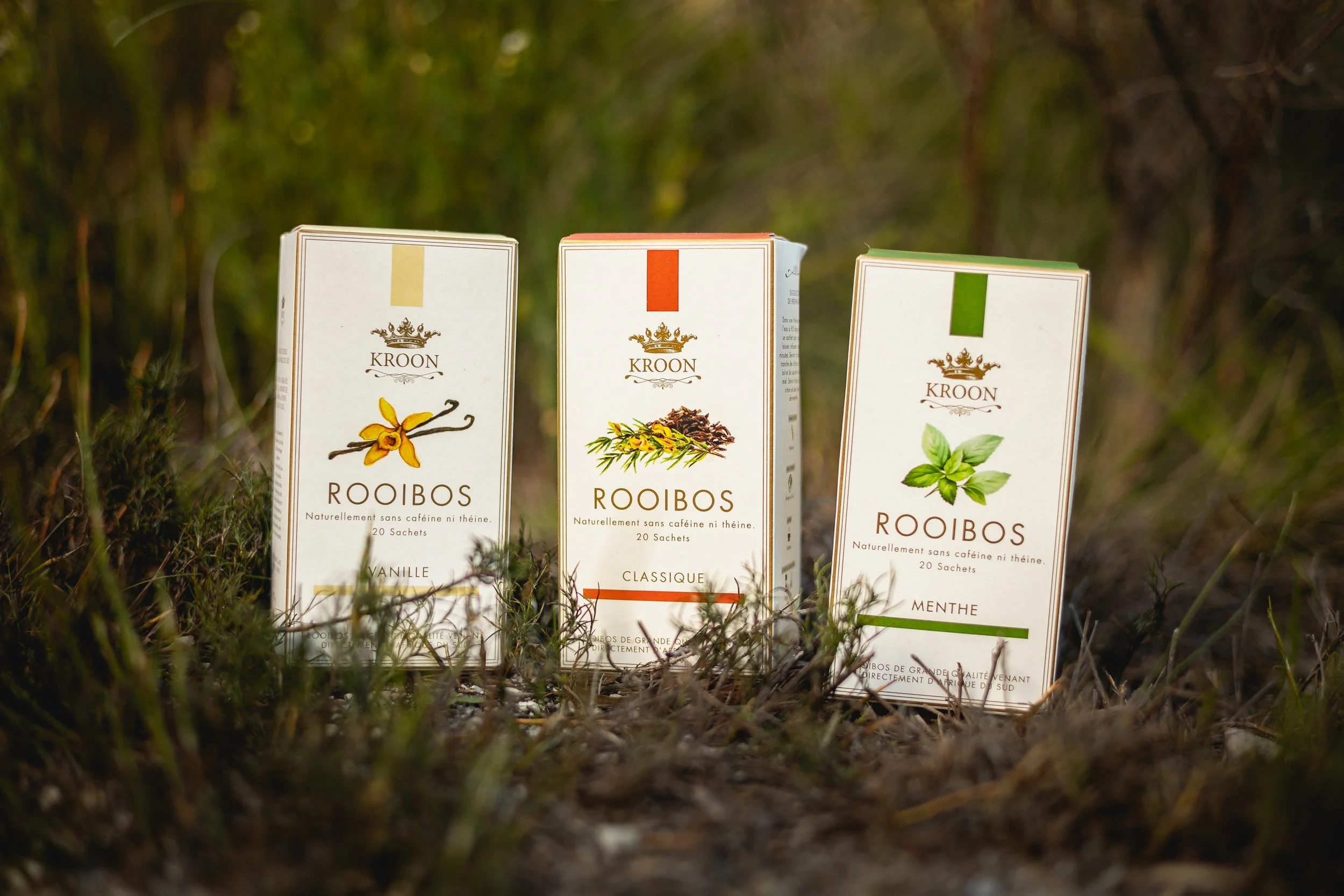 Trois paquets de rooibos se tenant sur le sol parmi des herbes, avec un fond de forêt. Le paquet de gauche est à la vanille, le centre est classique, et celui de droite à la menthe.