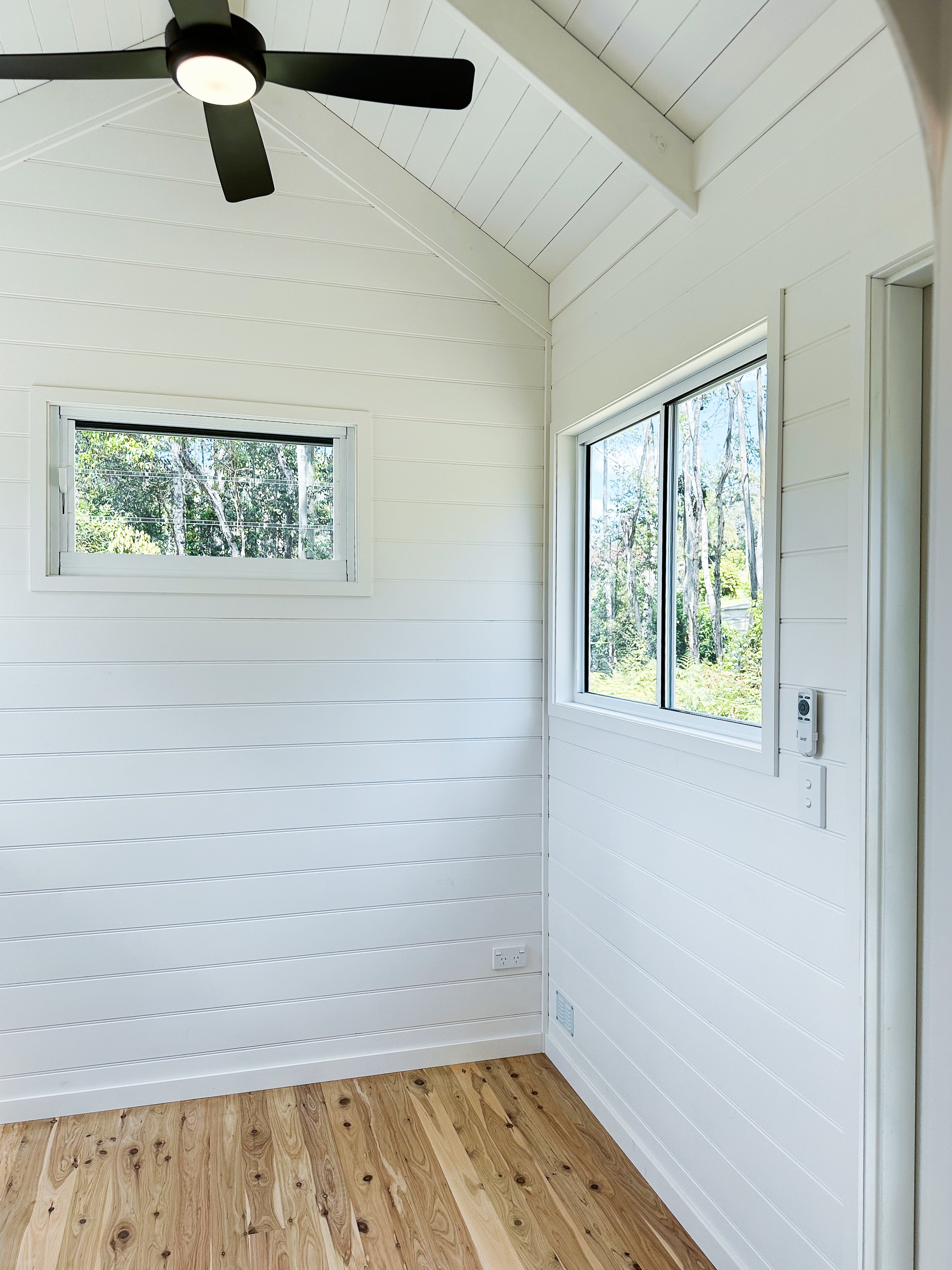Minimalist small room with white shiplap walls, light wood flooring, black ceiling fan, and large windows.