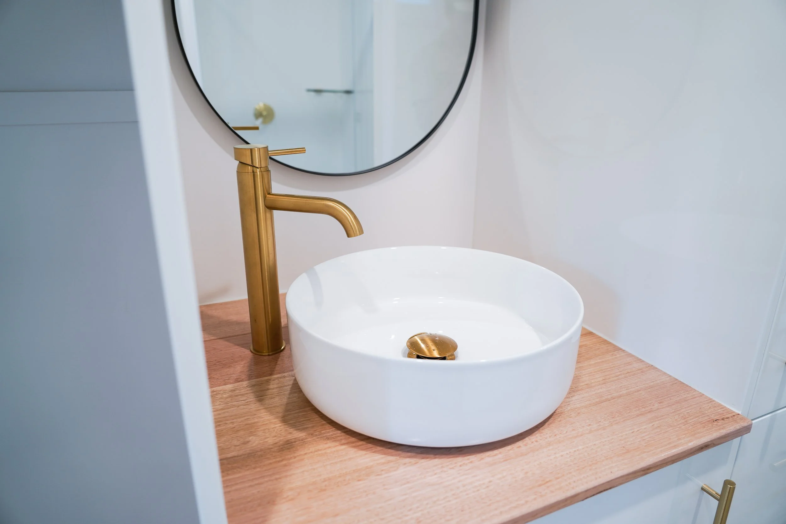 Modern bathroom sink with a white round vessel sink, gold faucet, and wooden countertop, featuring a round mirror in the background.