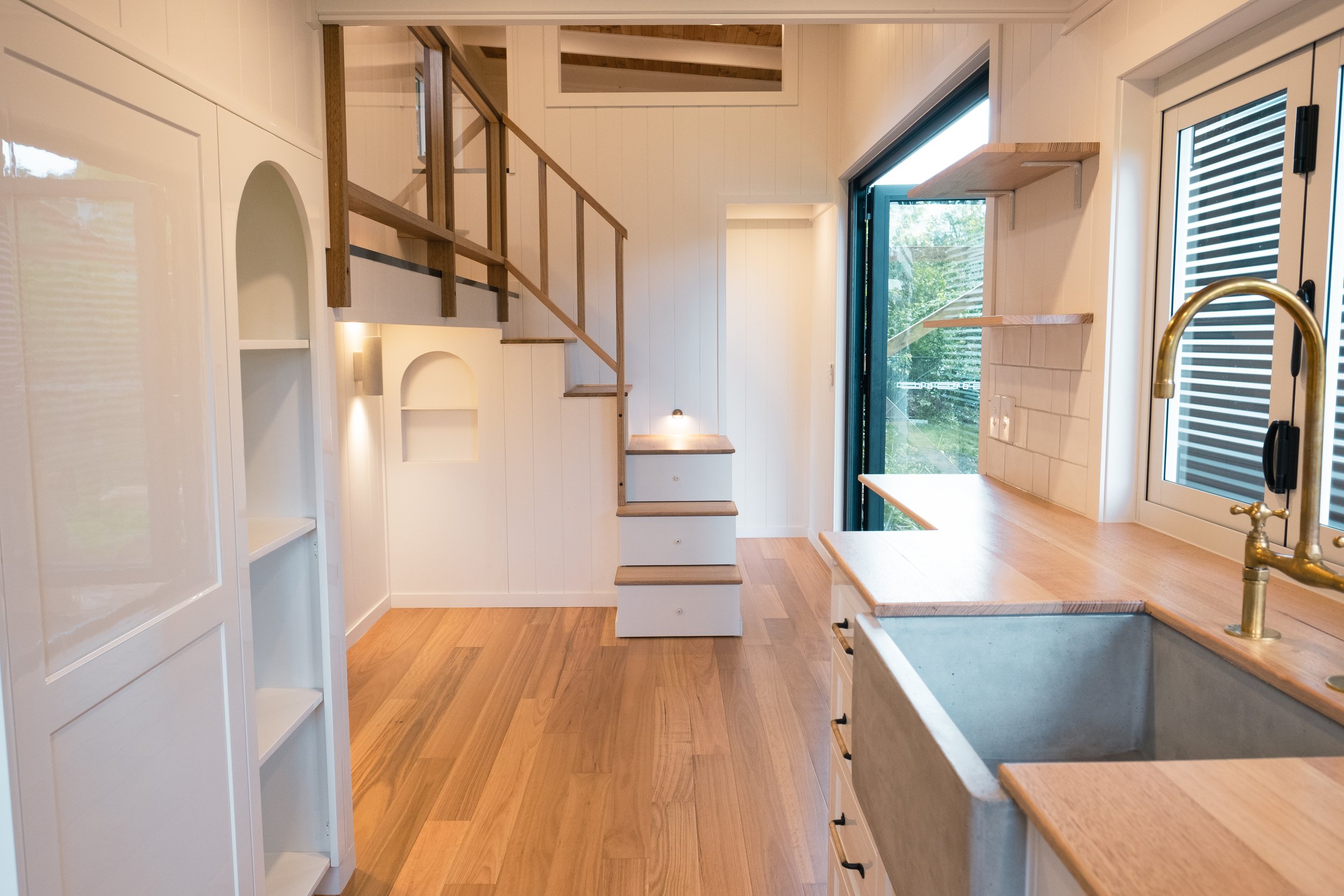 Interior of a small, modern tiny house with light wood floors, white walls, and minimalist design. Features include a staircase with built-in storage, a kitchen area with wooden countertops, a large farmhouse sink, open shelves, and a window with a v