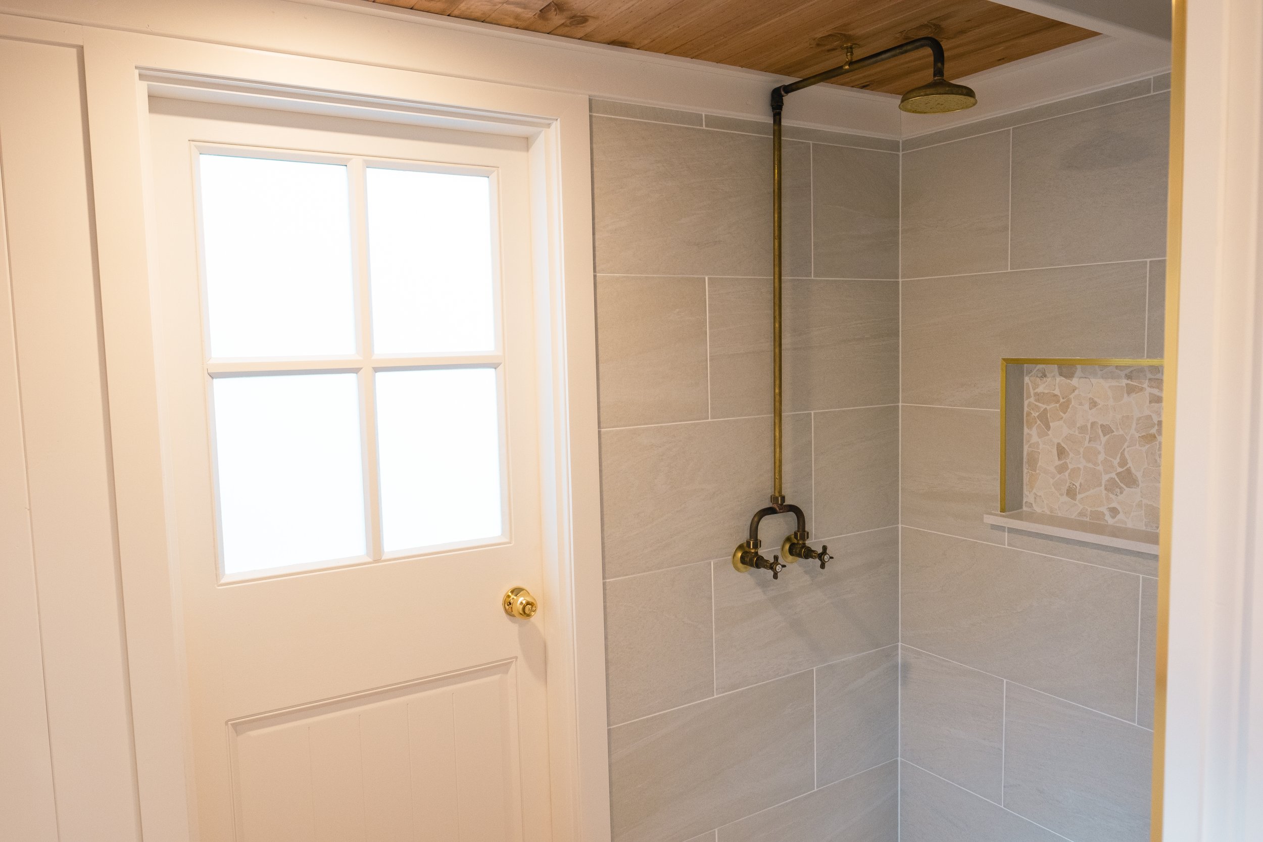 Modern bathroom shower with brass fixtures, beige tiles, showerhead, built-in shelf, and a white door with a frosted window.