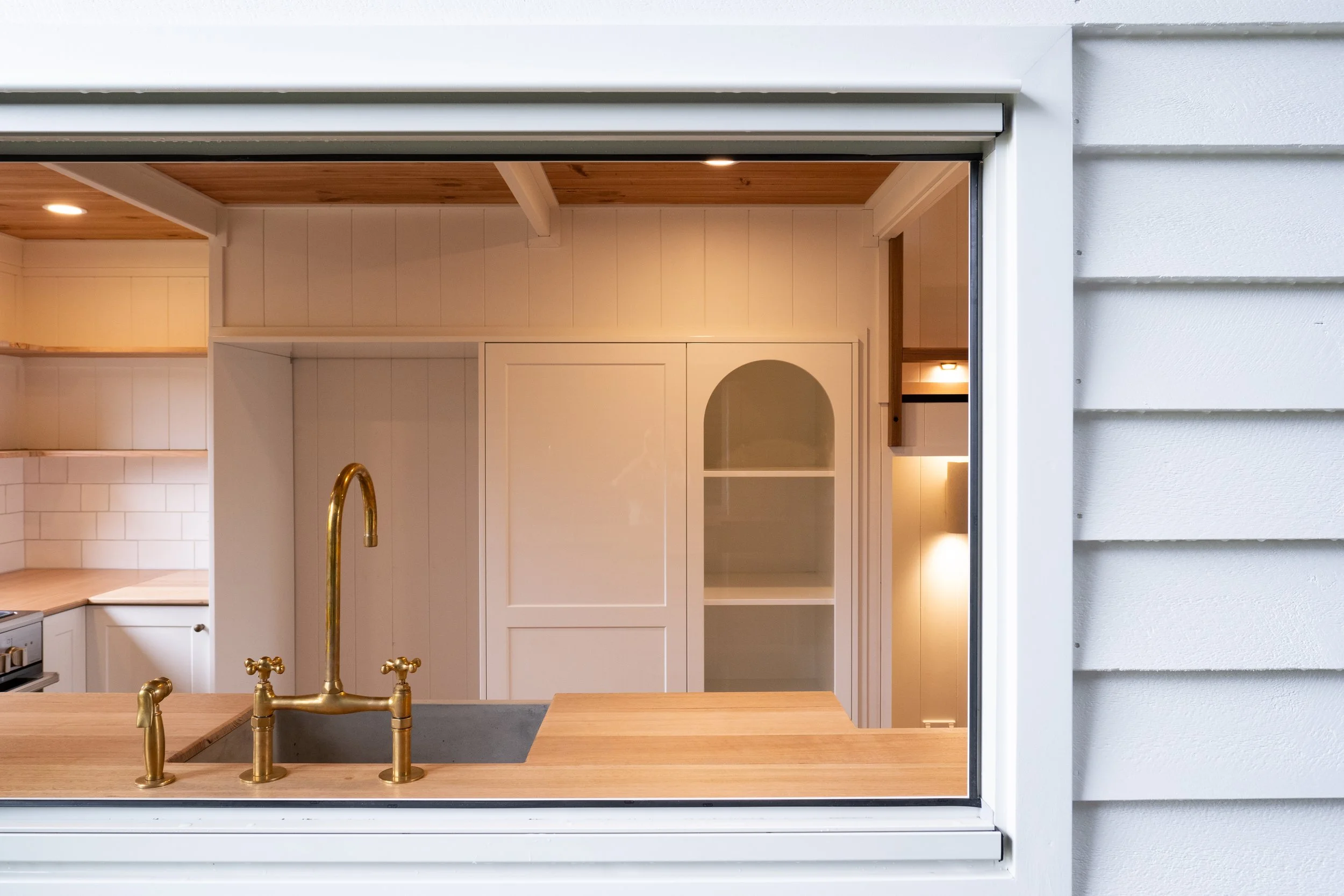 Modern kitchen interior seen through a window, featuring a gold faucet, wooden countertop, white cabinetry, and tiled backsplash.